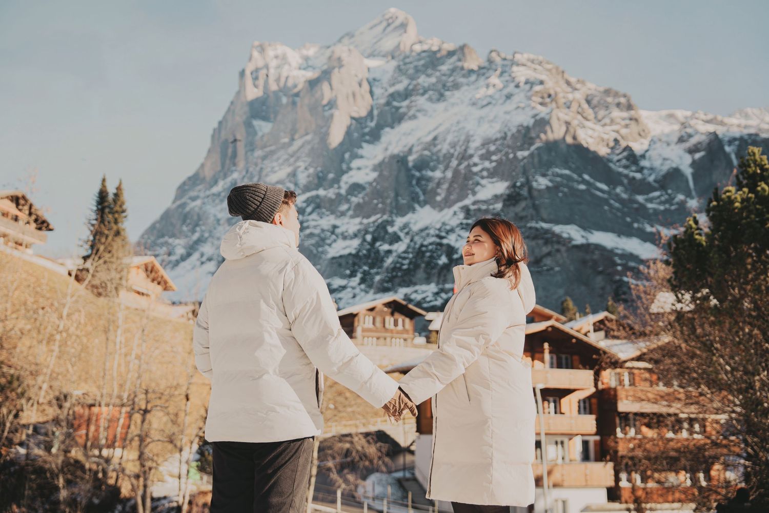 Couple holding hands facing each other with dramatic snow-capped mountain and alpine village behind.