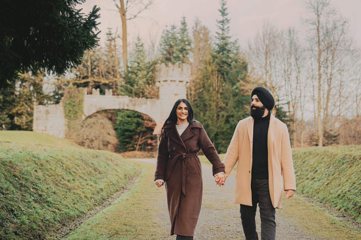 Couple holding hands and strolling along a path with a stone castle bridge in the background.