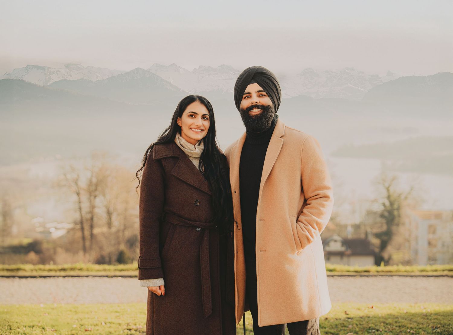 Sikh couple smiling together outdoors with misty mountain range visible in the background.