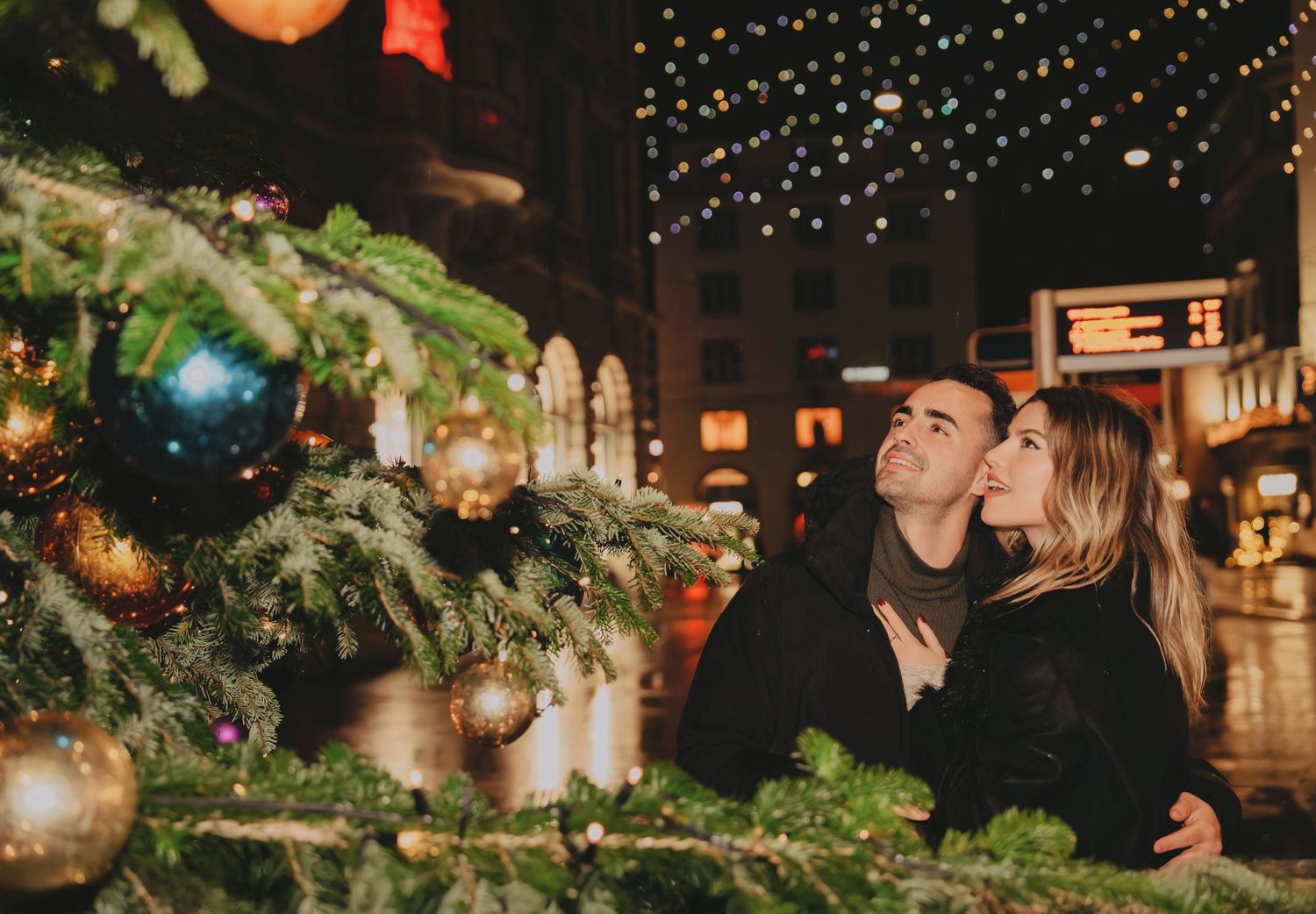Romantic couple gazing up at a decorated Christmas tree with ornaments at night.