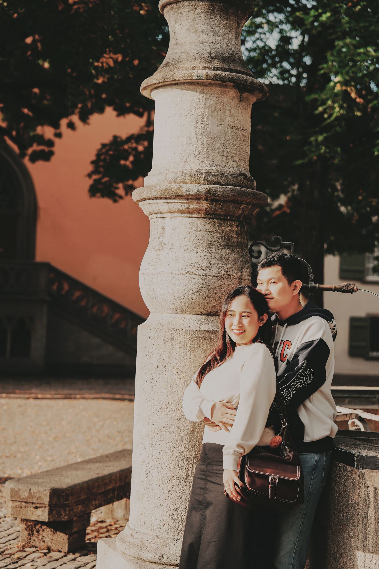 Couple posing by a stone column in a European courtyard, woman smiling in white sweater with brown crossbody bag.