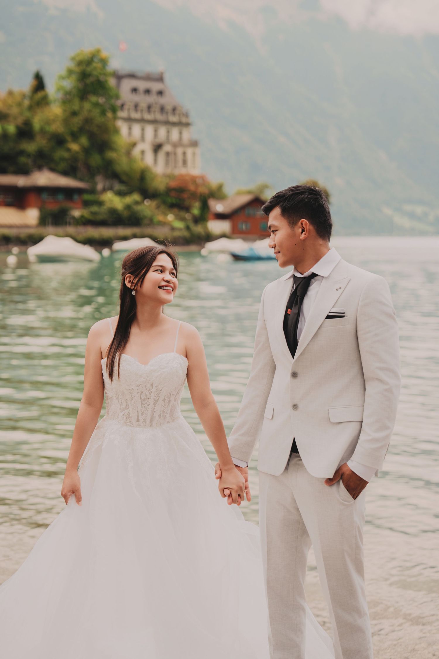 Smiling bride and groom holding hands and looking at each other by a stunning alpine lake backdrop.