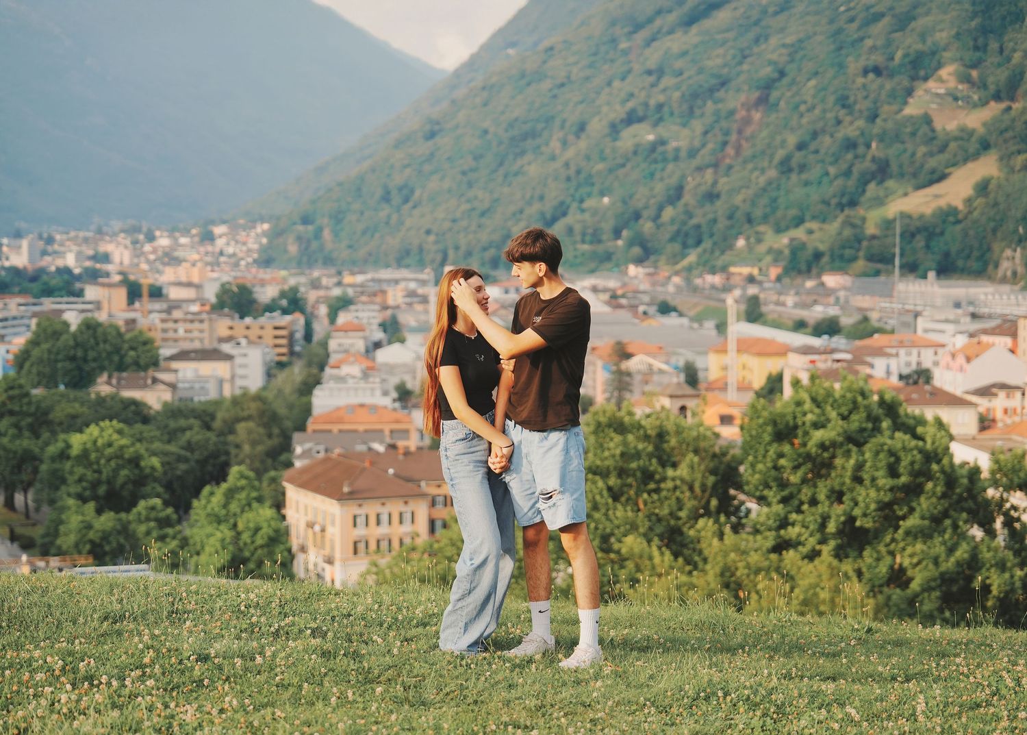 Romantic couple embracing on green hillside with Italian mountain town panorama behind them.