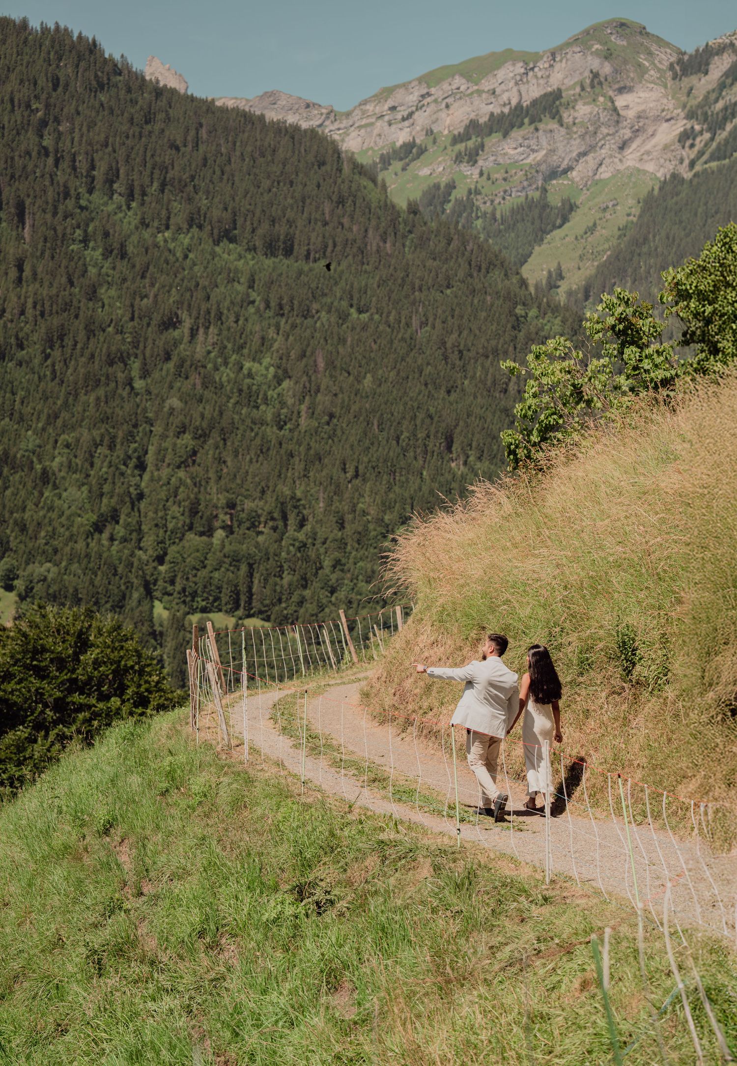 Newlywed couple walking hand in hand along a winding mountain path surrounded by green Alpine scenery.