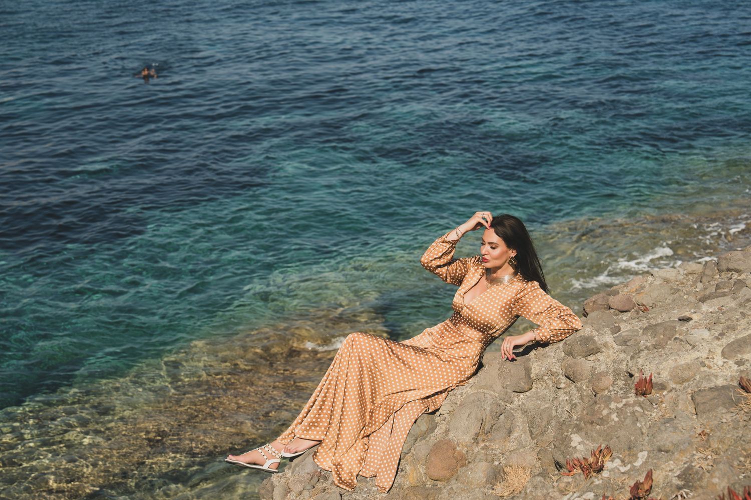 Woman in beige maxi dress posing on rocky coastline with turquoise sea water in background.