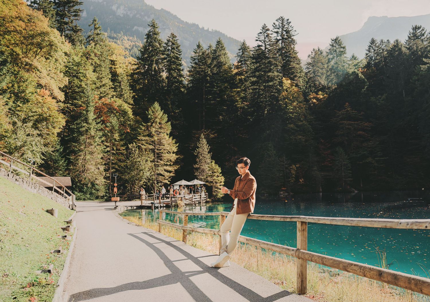 Man standing by turquoise alpine lake on a sunny autumn day with forested mountains in background.