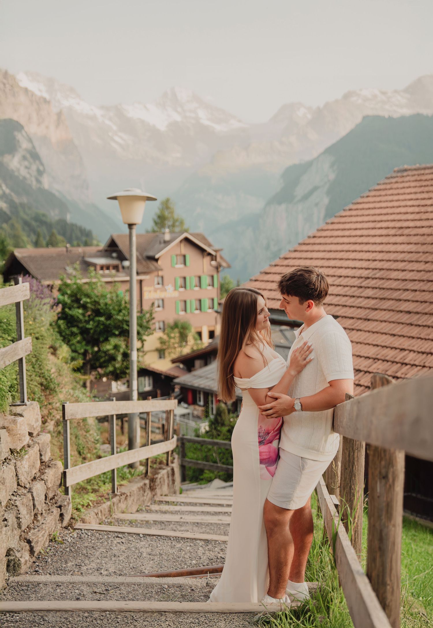 Romantic couple embracing on stone staircase in charming Swiss Alpine village with dramatic mountain backdrop.