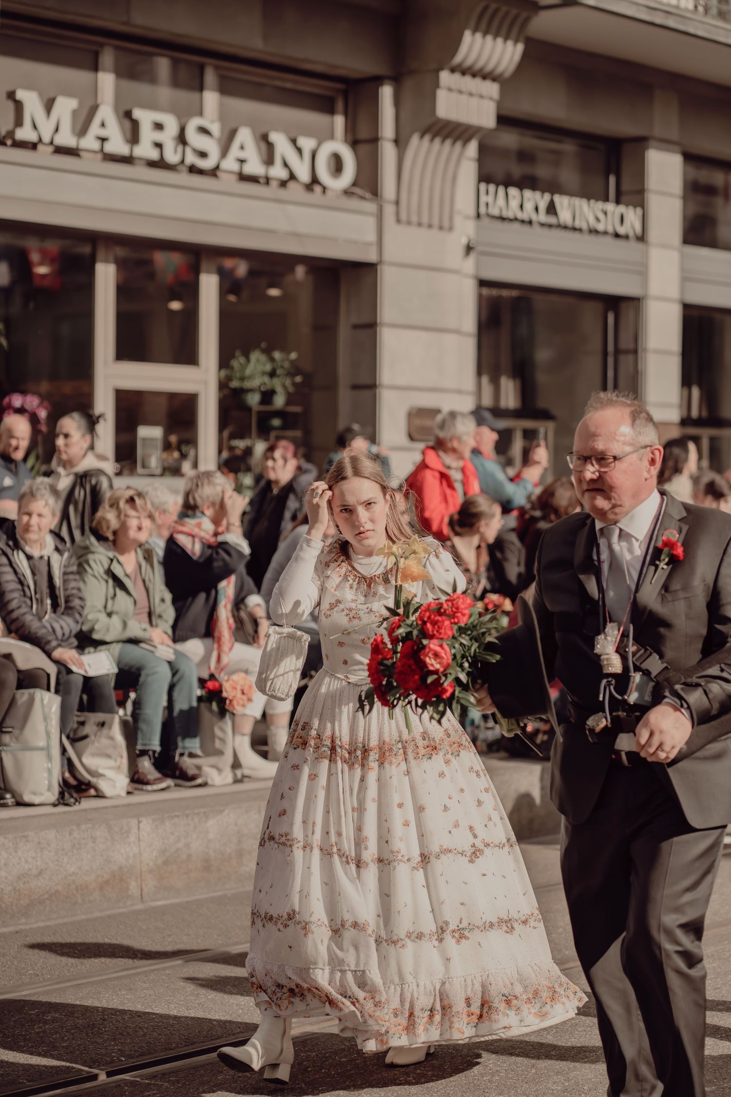 Woman in an embroidered vintage dress carries red roses while walking in a festive street parade.