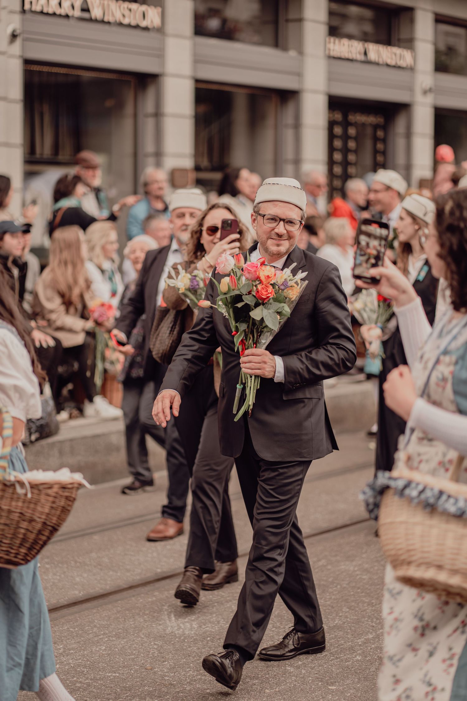 Smiling man in dark suit carries colorful rose bouquet while walking through a festive street parade.