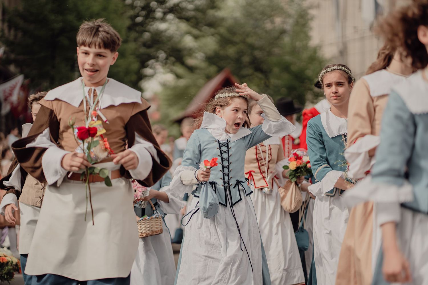 Young participants in historical costumes carry flowers and paper decorations in a lively street parade.