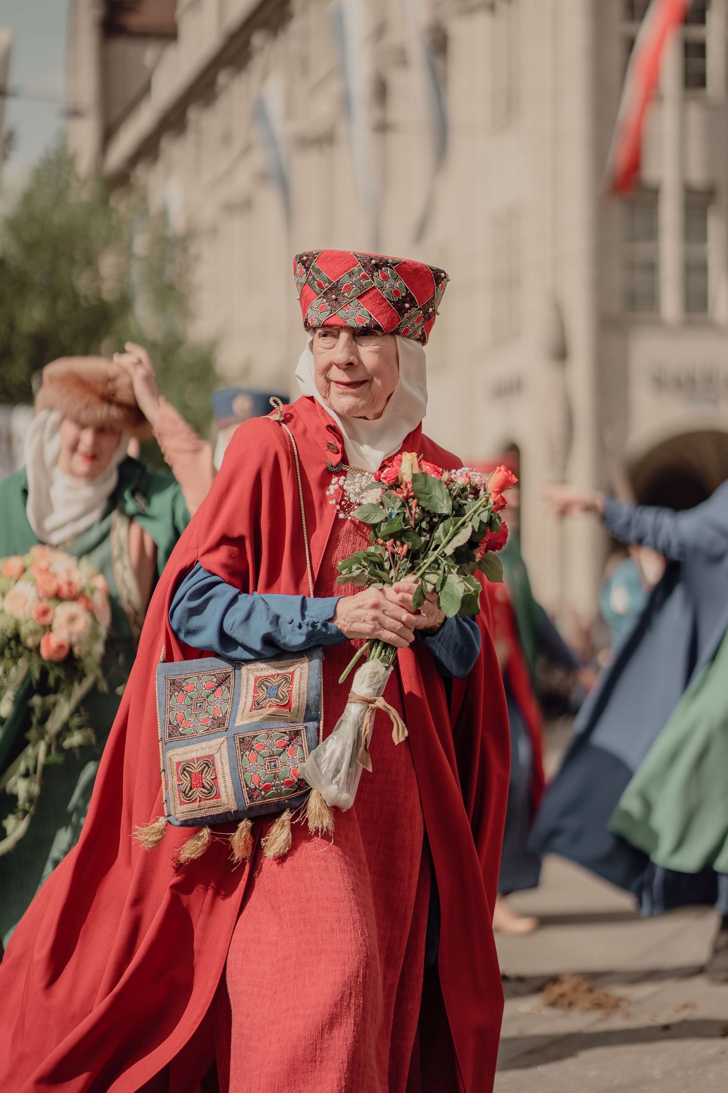 Woman in red medieval costume with embroidered hat holds rose bouquet during historical street parade.