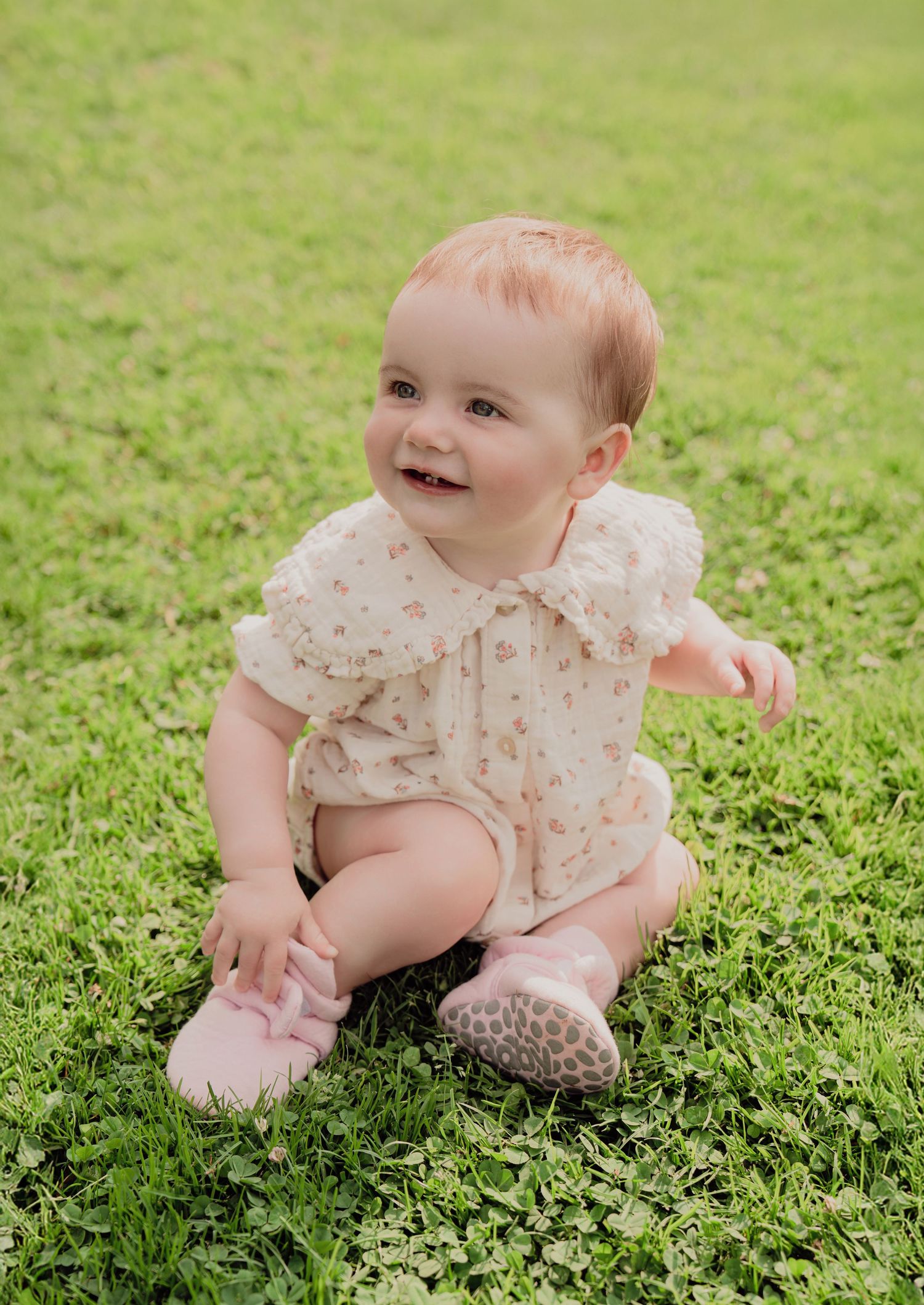 Adorable baby in floral romper sitting on green grass smiling, wearing pink shoes.