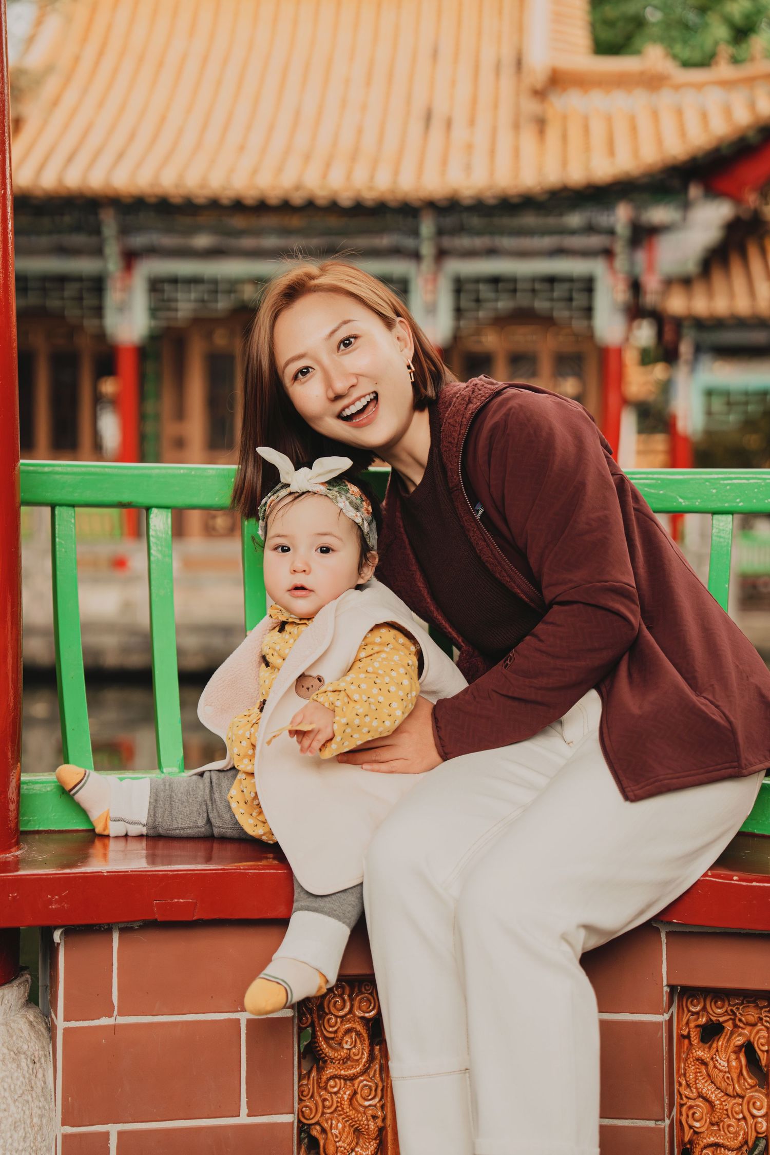 Young Asian mother sitting with cute baby in yellow outfit at ornate Chinese temple railing.