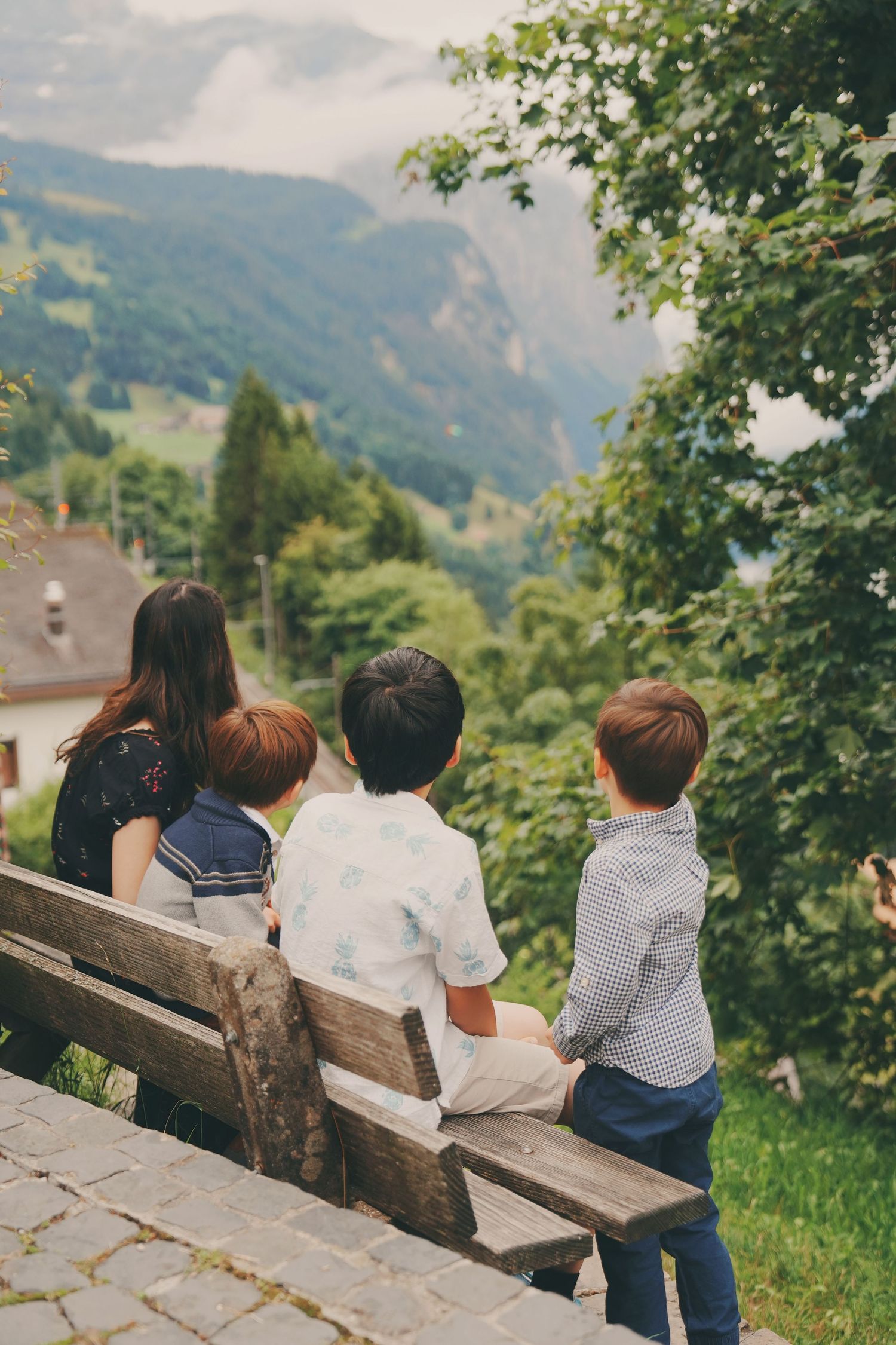 A mother and three children sit on a wooden bench overlooking a lush Swiss alpine valley.