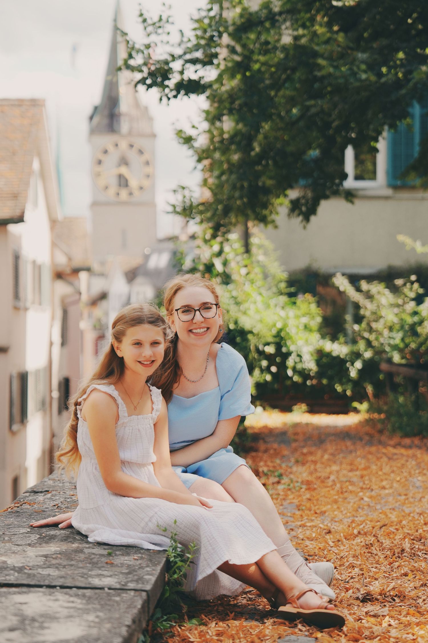 A woman in a blue dress and a girl in white sit together on a stone wall with a church tower backdrop.