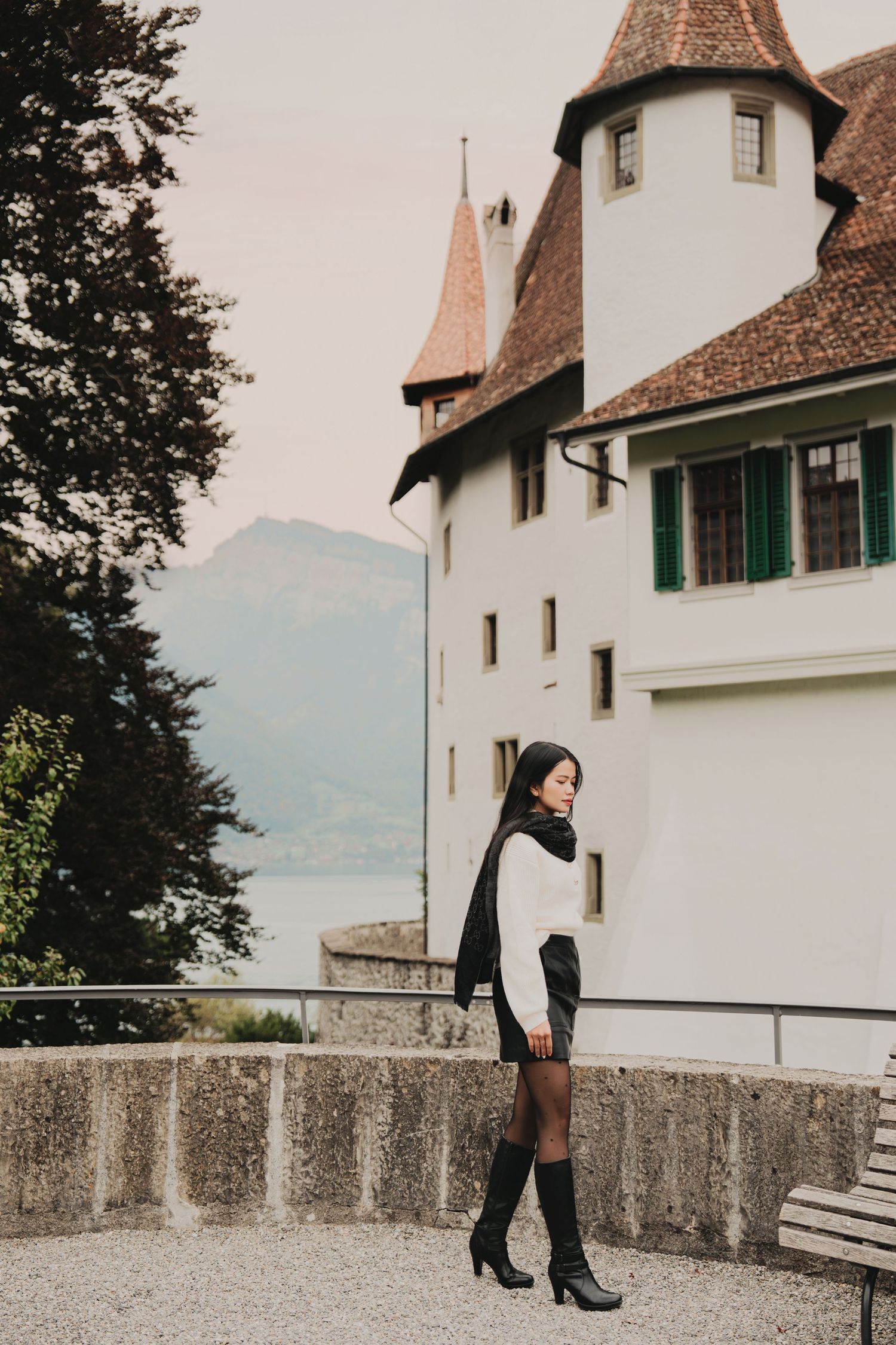 Woman in white sweater and black skirt stands beside historic white Swiss castle with mountain view.