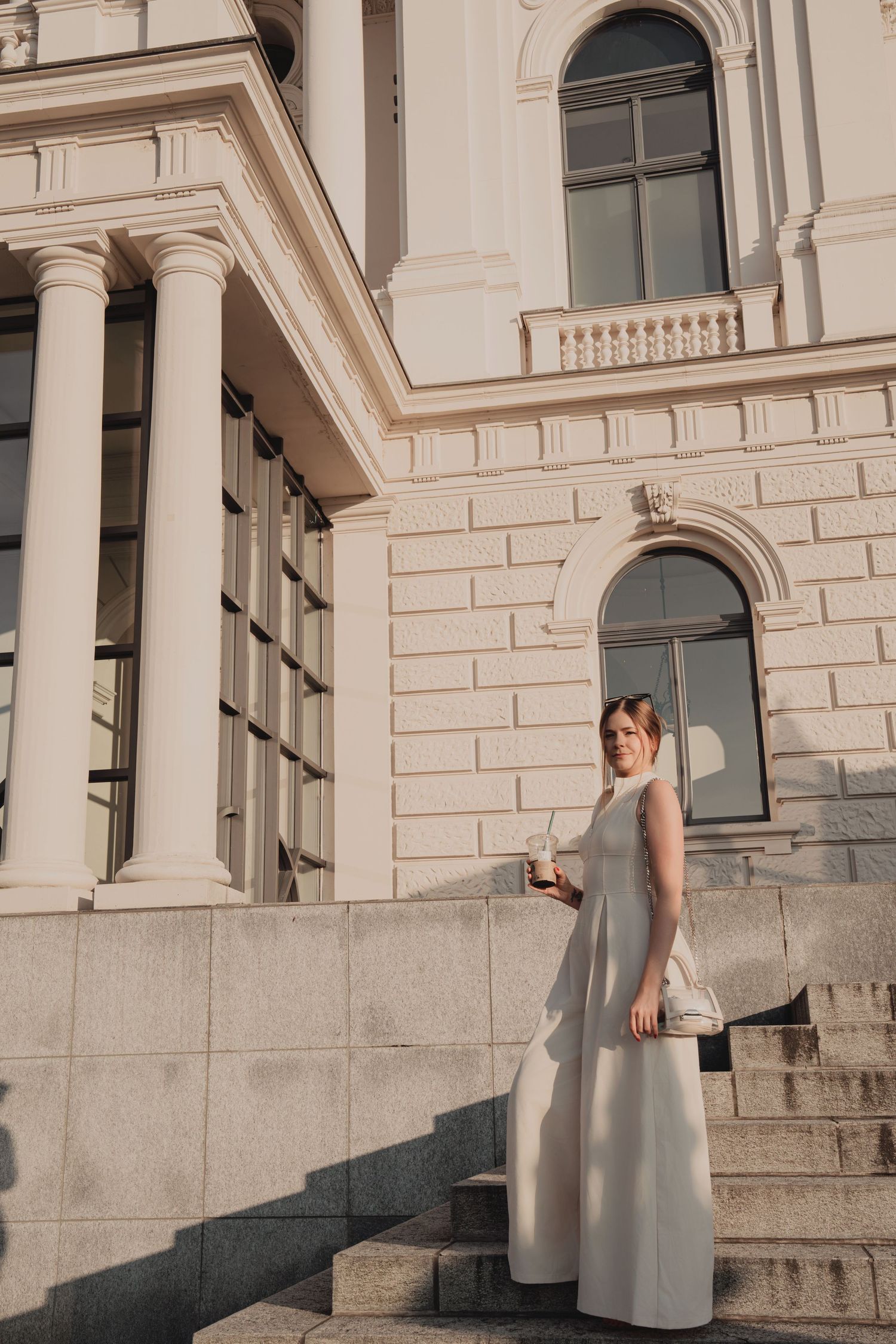 Elegant woman in white maxi dress standing on steps of a grand neoclassical building.