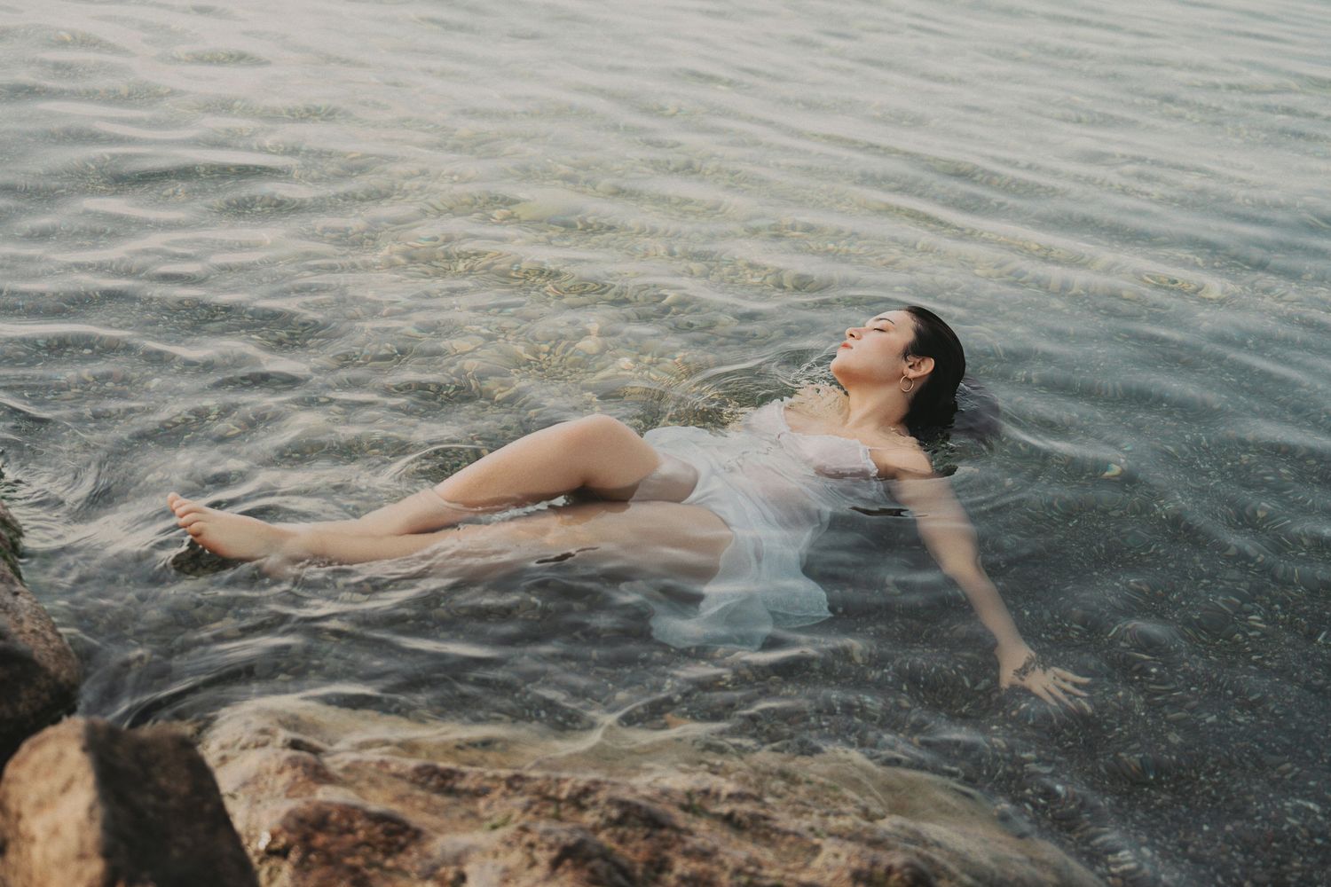 Woman in sheer white dress floating in shallow ocean water near rocky shore at dusk.