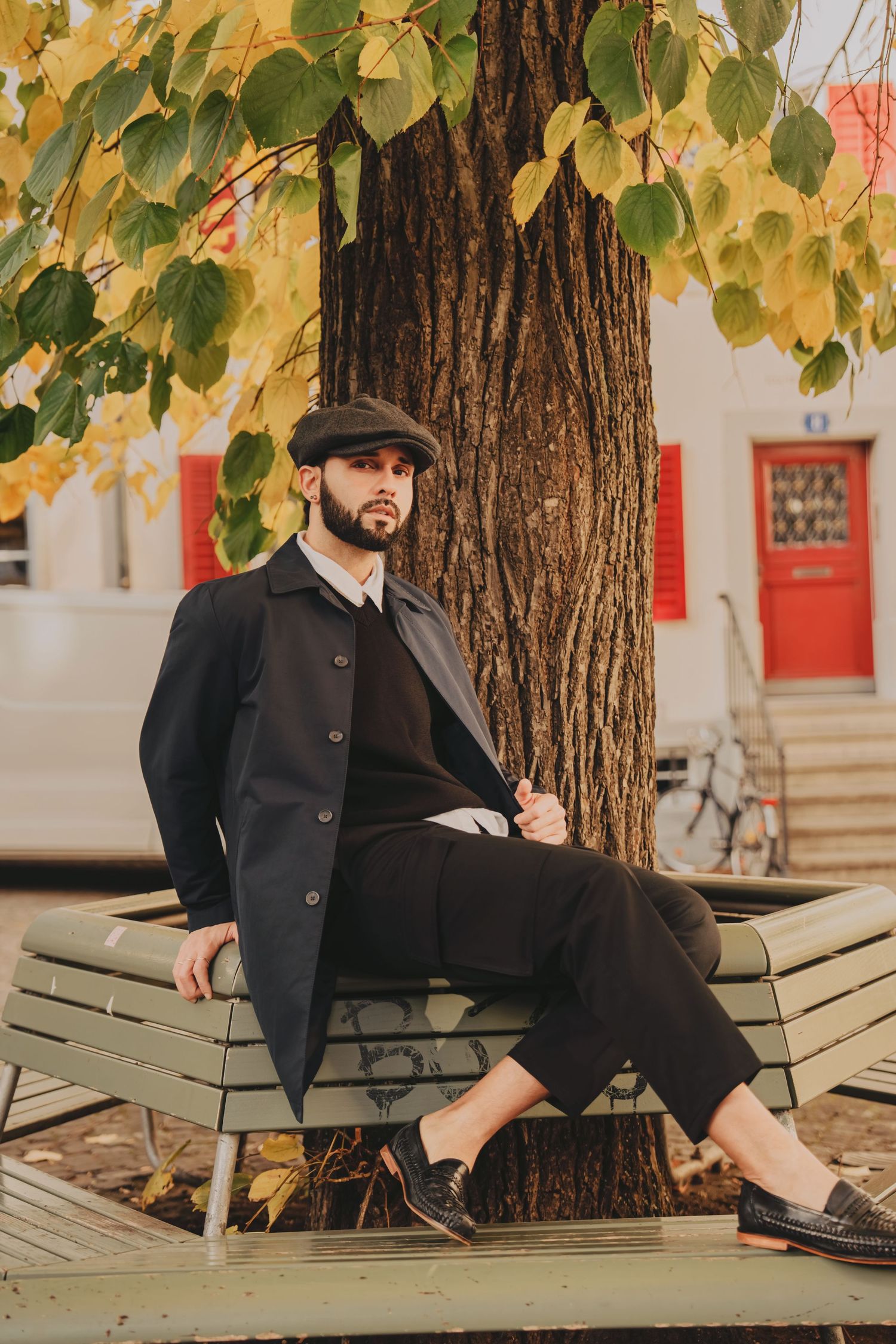 Stylish man in dark coat and flat cap sitting on bench under large tree with autumn foliage.