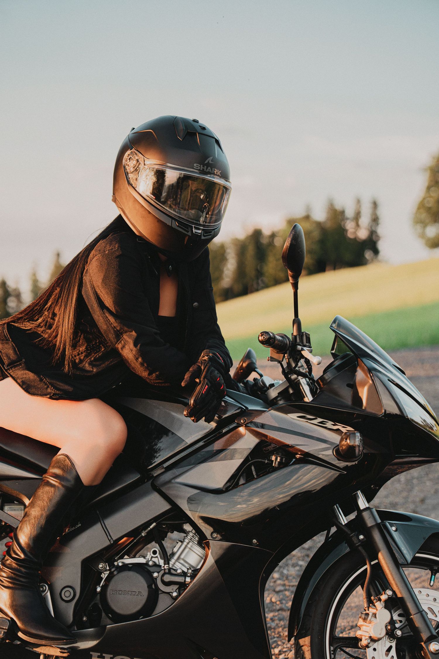 Woman in a black helmet and outfit sitting on a black sport motorcycle in a sunny rural setting.