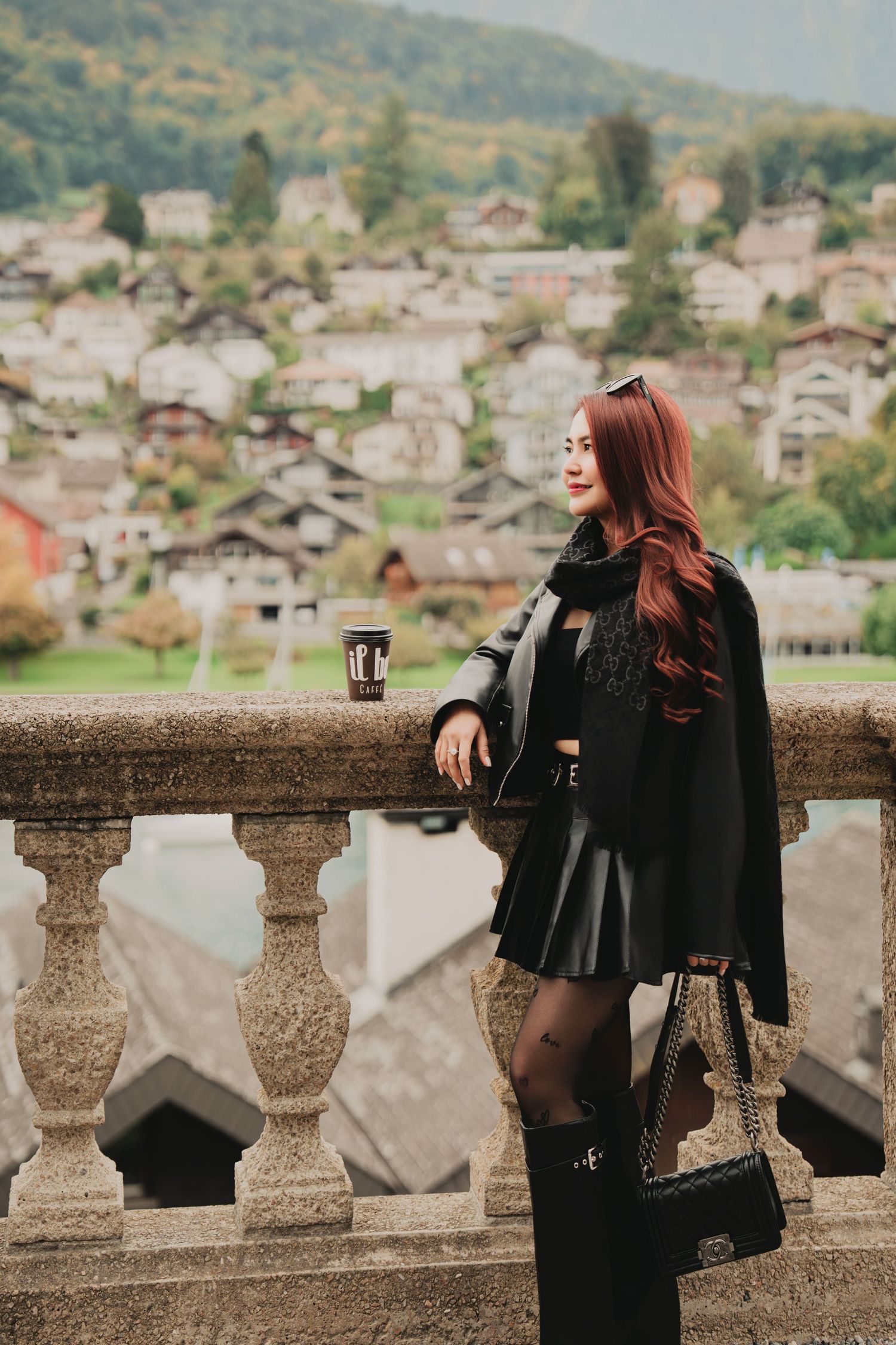 Red-haired woman in black leather mini skirt poses at stone balcony with Swiss mountain village behind.