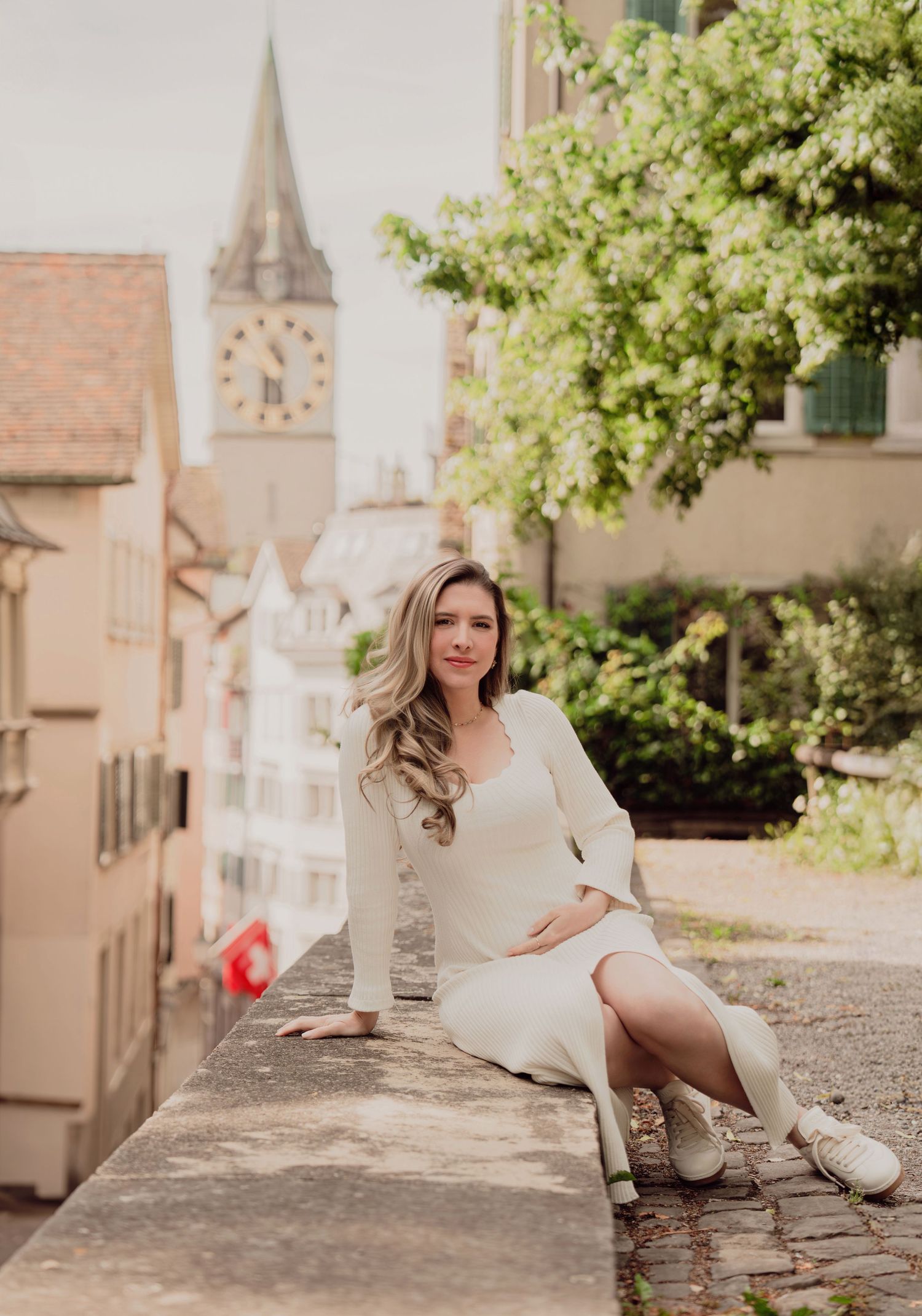 Pregnant woman in elegant white dress sits on stone wall in sunny Zurich old town with church tower behind.