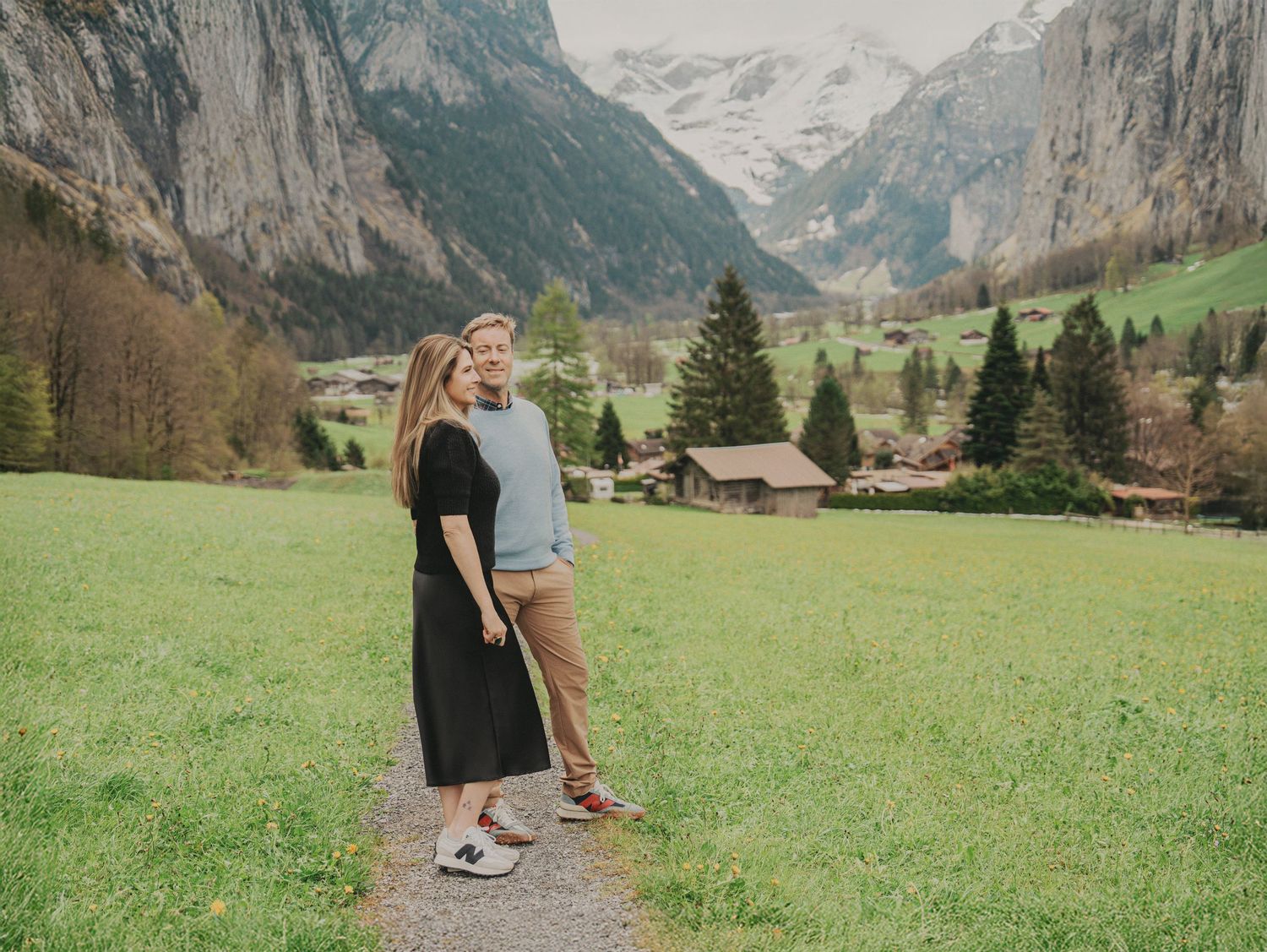 Couple standing together on alpine trail with picturesque Swiss mountain village and peaks behind.