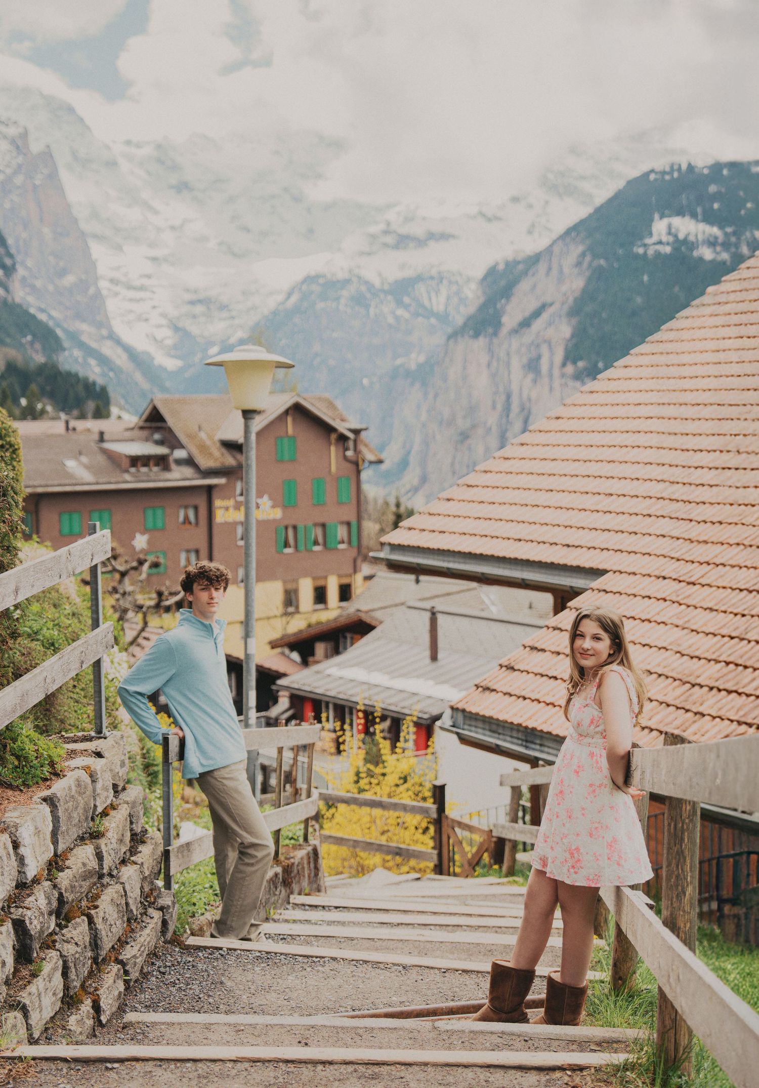 Young couple posing on wooden staircase in scenic Swiss Alpine village with snow-capped mountain views.