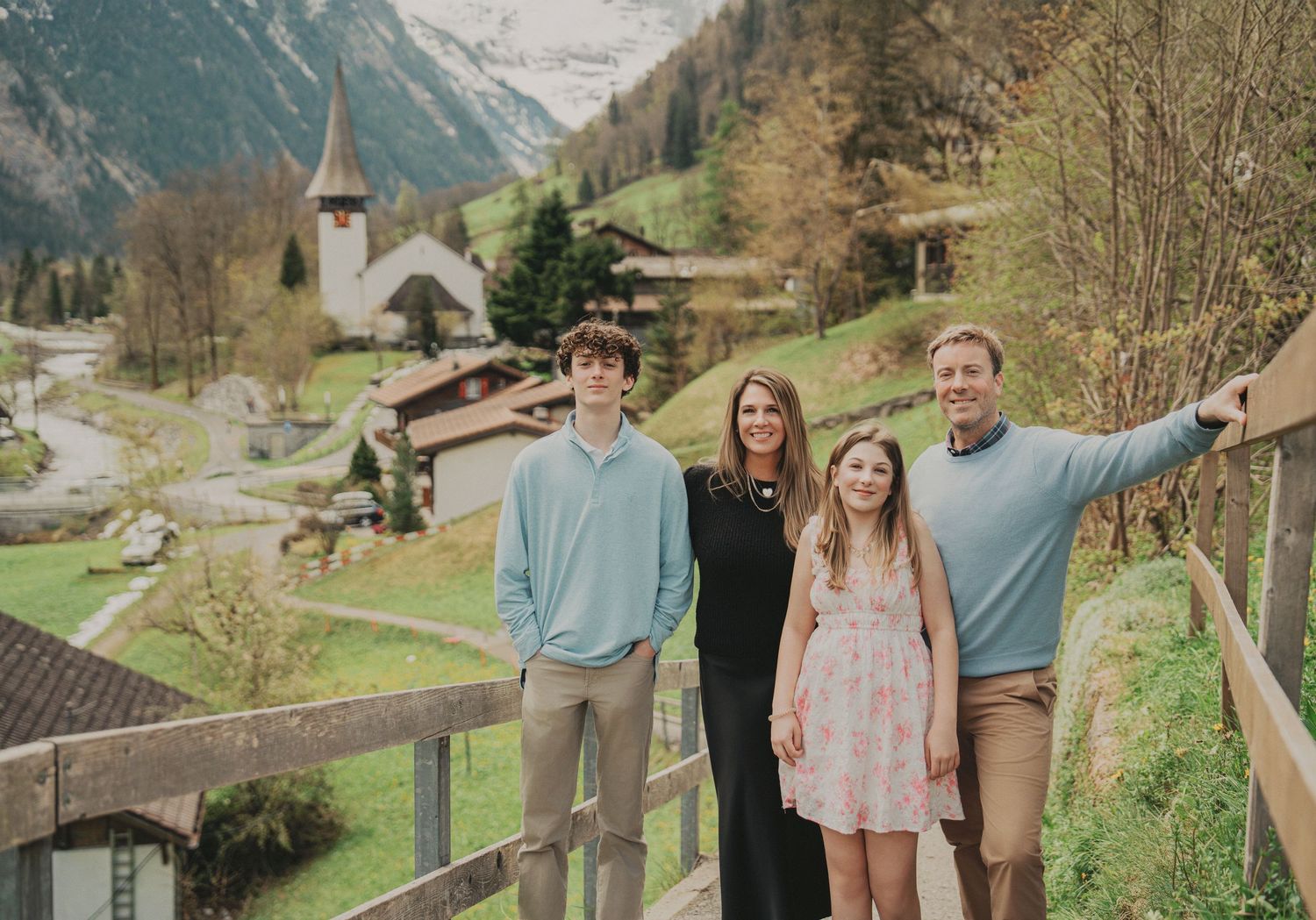 Family of four posing on a wooden bridge with a Swiss alpine village and snow-capped mountains in the background.