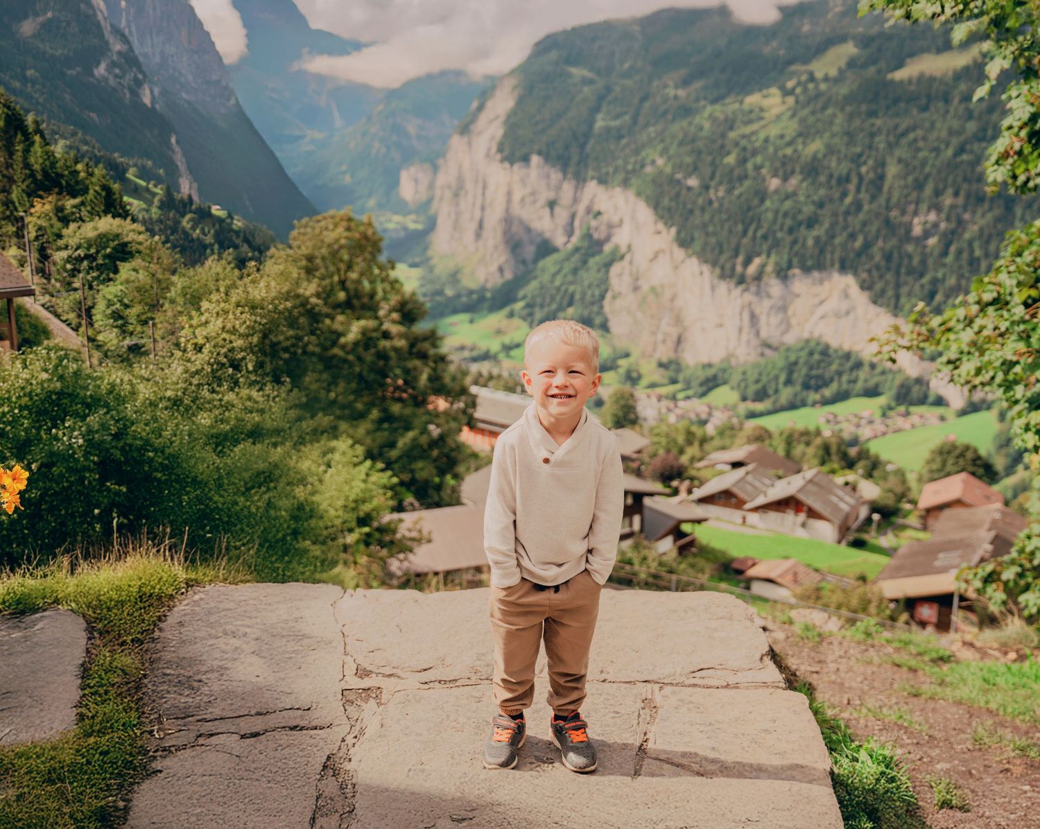 Happy young blonde boy in beige sweater standing on stone ledge with lush Alpine valley behind him.