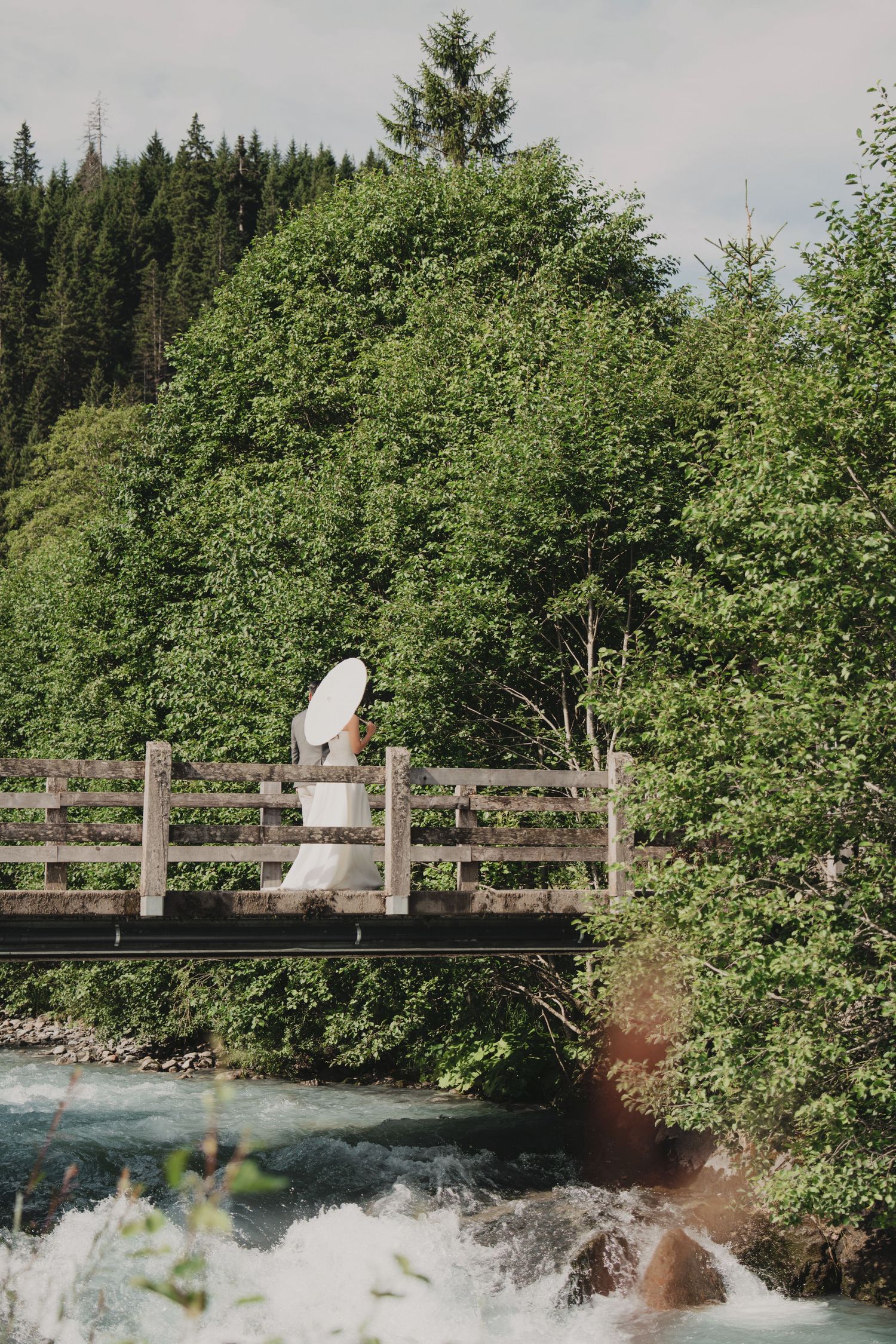 Bride in white gown holding parasol walks across rustic wooden bridge over turquoise mountain river.