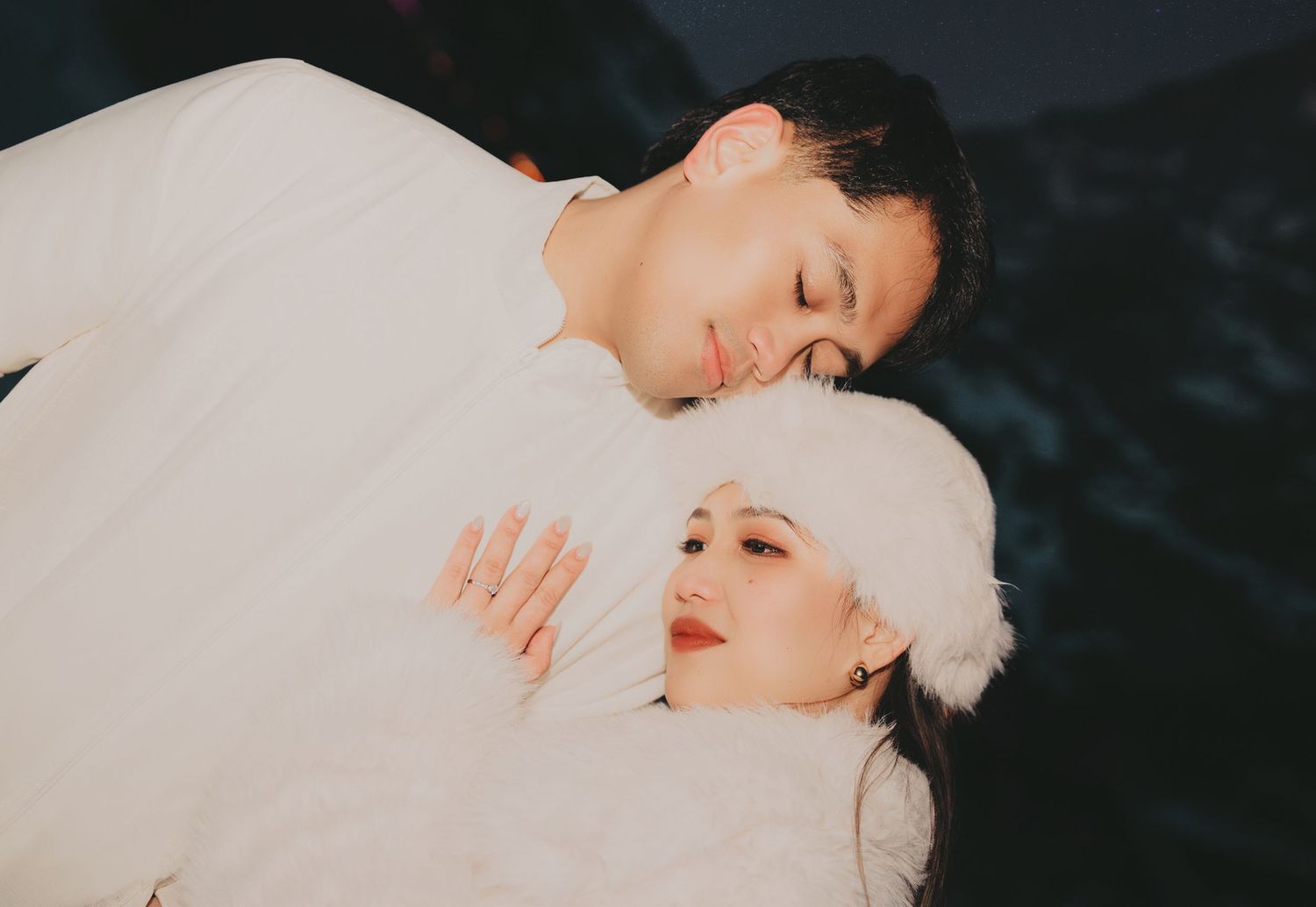 Couple in white winter attire cuddling closely with eyes closed in snowy setting.