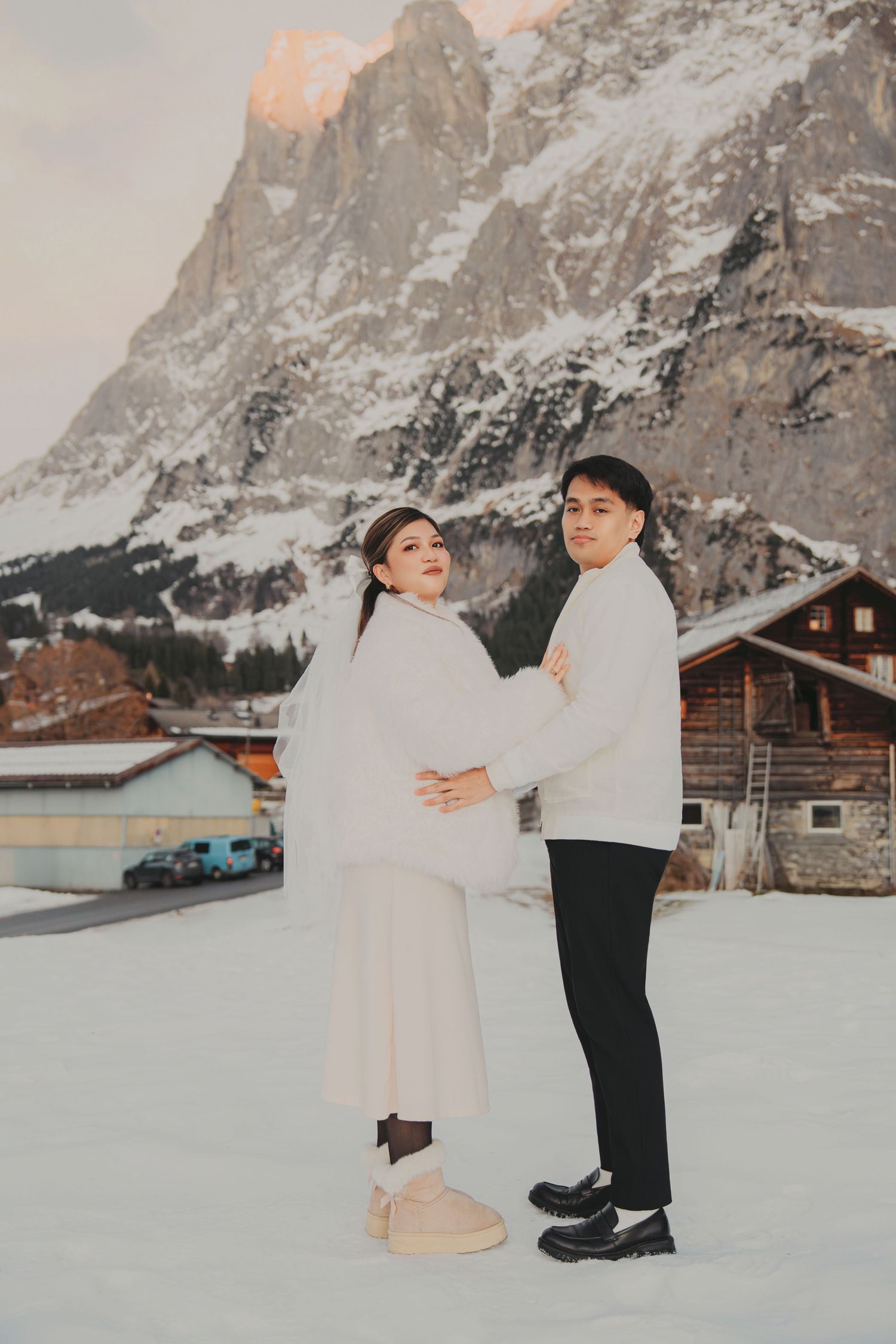 Couple in white winter outfits posing back-to-front on snow with dramatic mountain backdrop at dusk.
