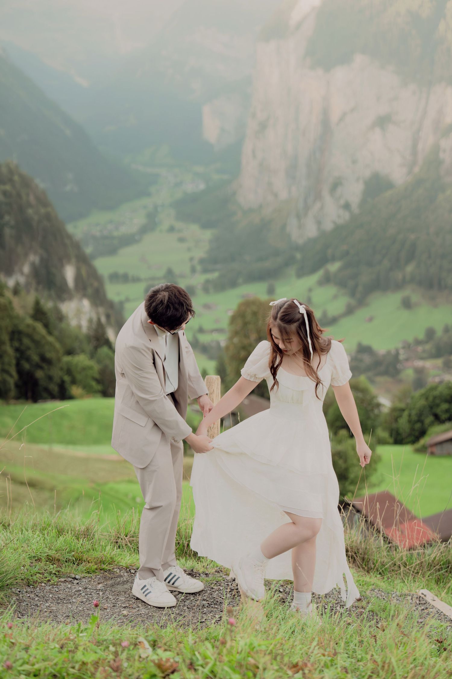 Couple dancing playfully on mountain path in Swiss Alps with green valley and rocky cliffs behind them.