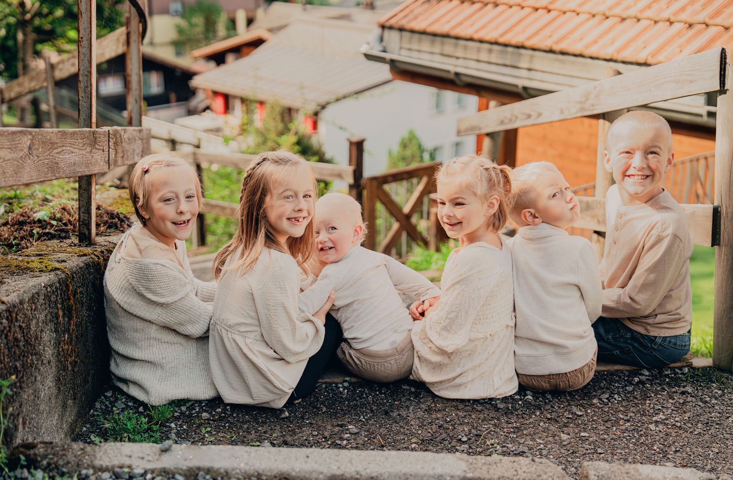 Six blonde children in matching cream outfits sitting together laughing on stone steps in a Swiss village.