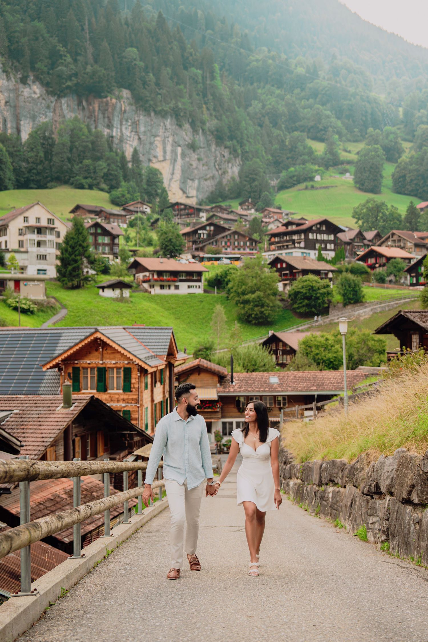 Couple walking hand in hand on a road through a charming Swiss Alpine village with mountain backdrop.