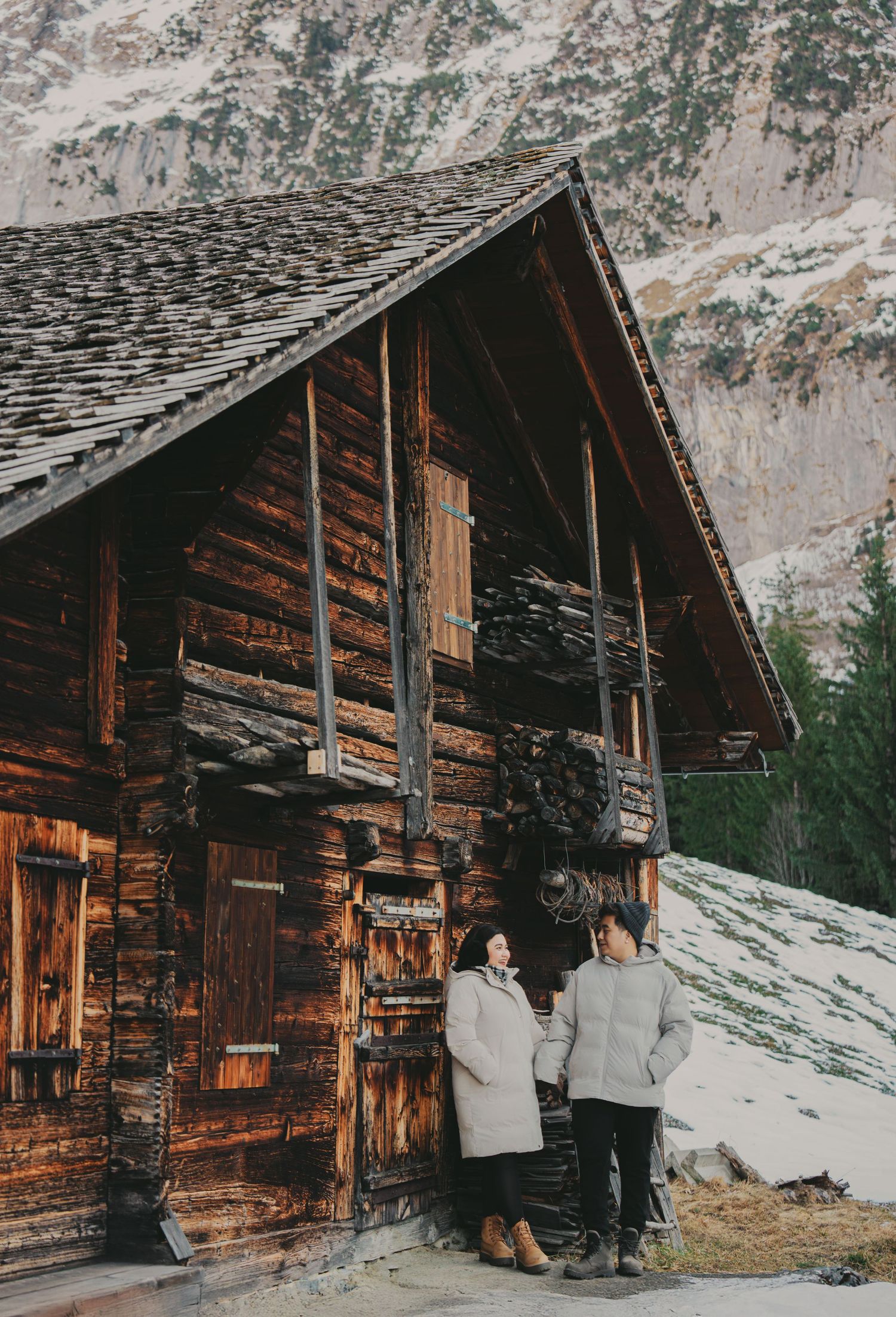 Couple in white winter coats holding hands beside weathered wooden mountain chalet in snowy Alps.