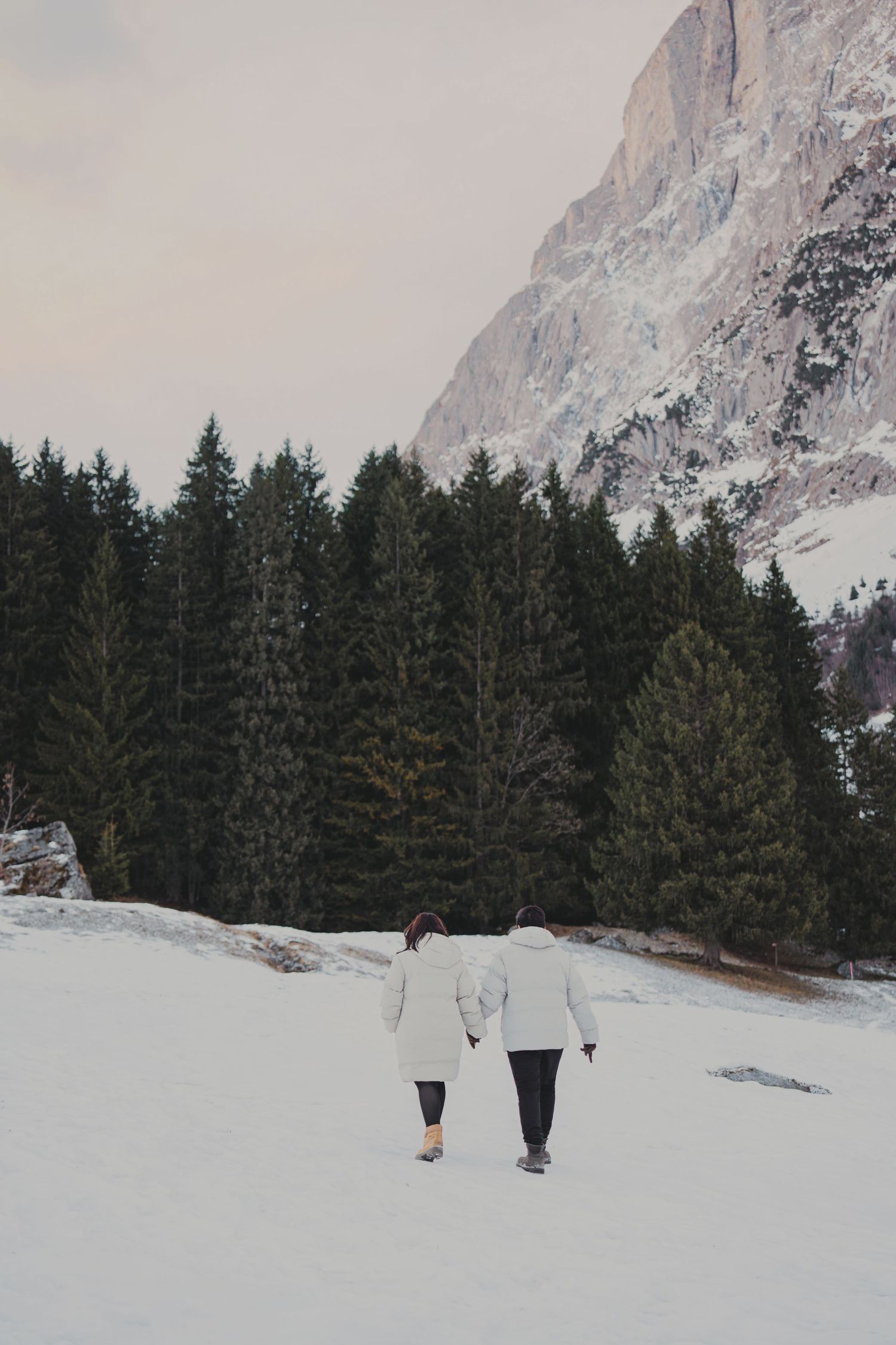 Couple holding hands walking toward pine forest across snowy field with rocky mountain towering above.