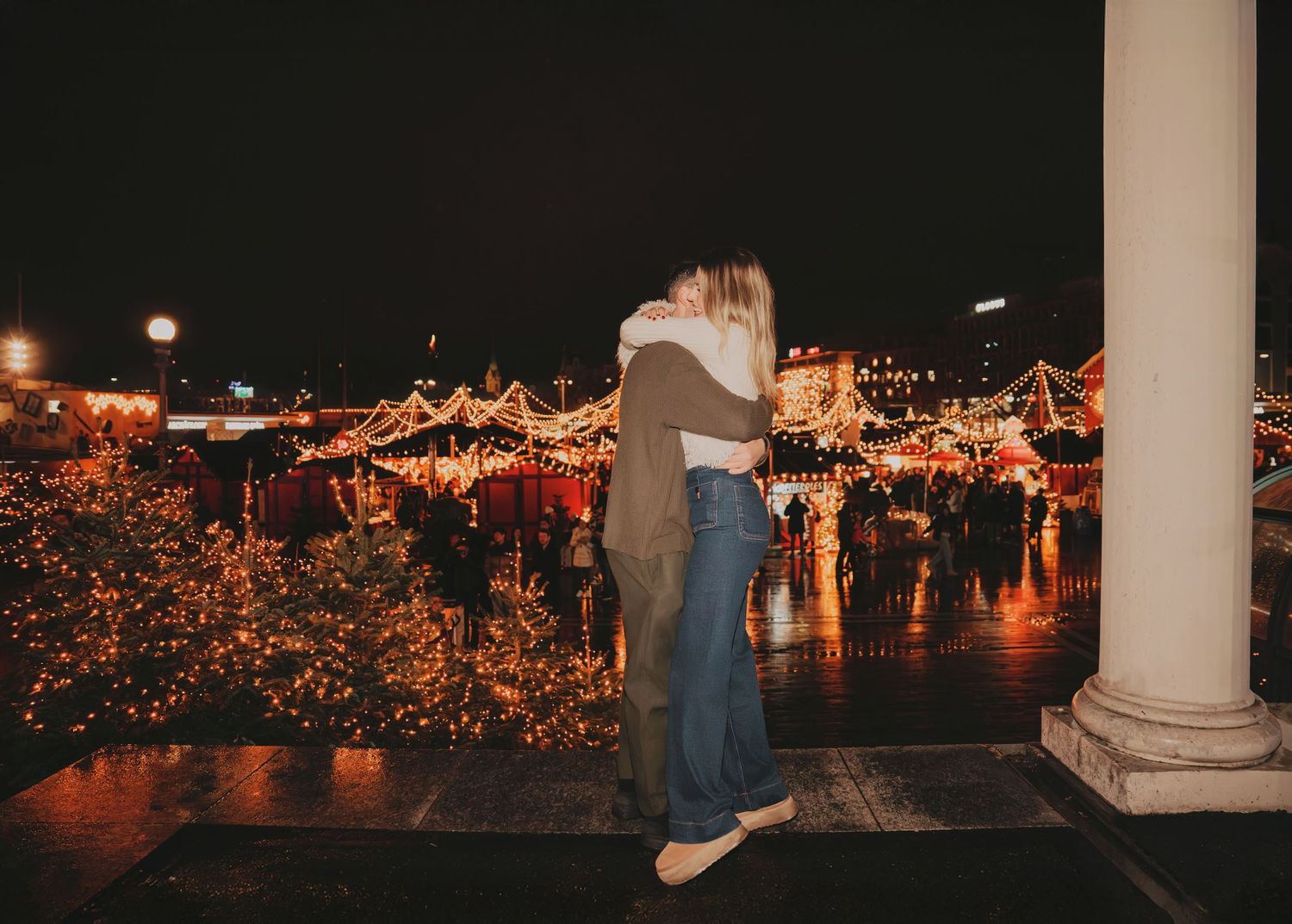 Couple embracing while overlooking a beautifully lit Christmas market with festive lights at night.