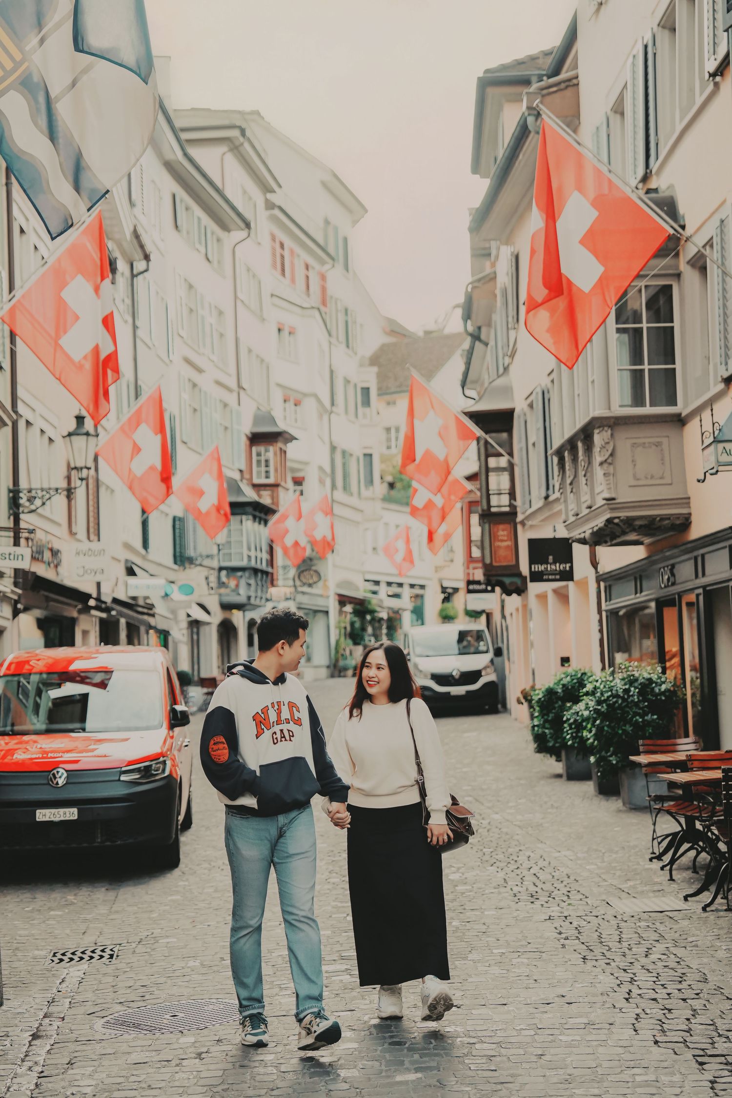 Couple holding hands and walking along a charming Swiss cobblestone street decorated with red Swiss flags.