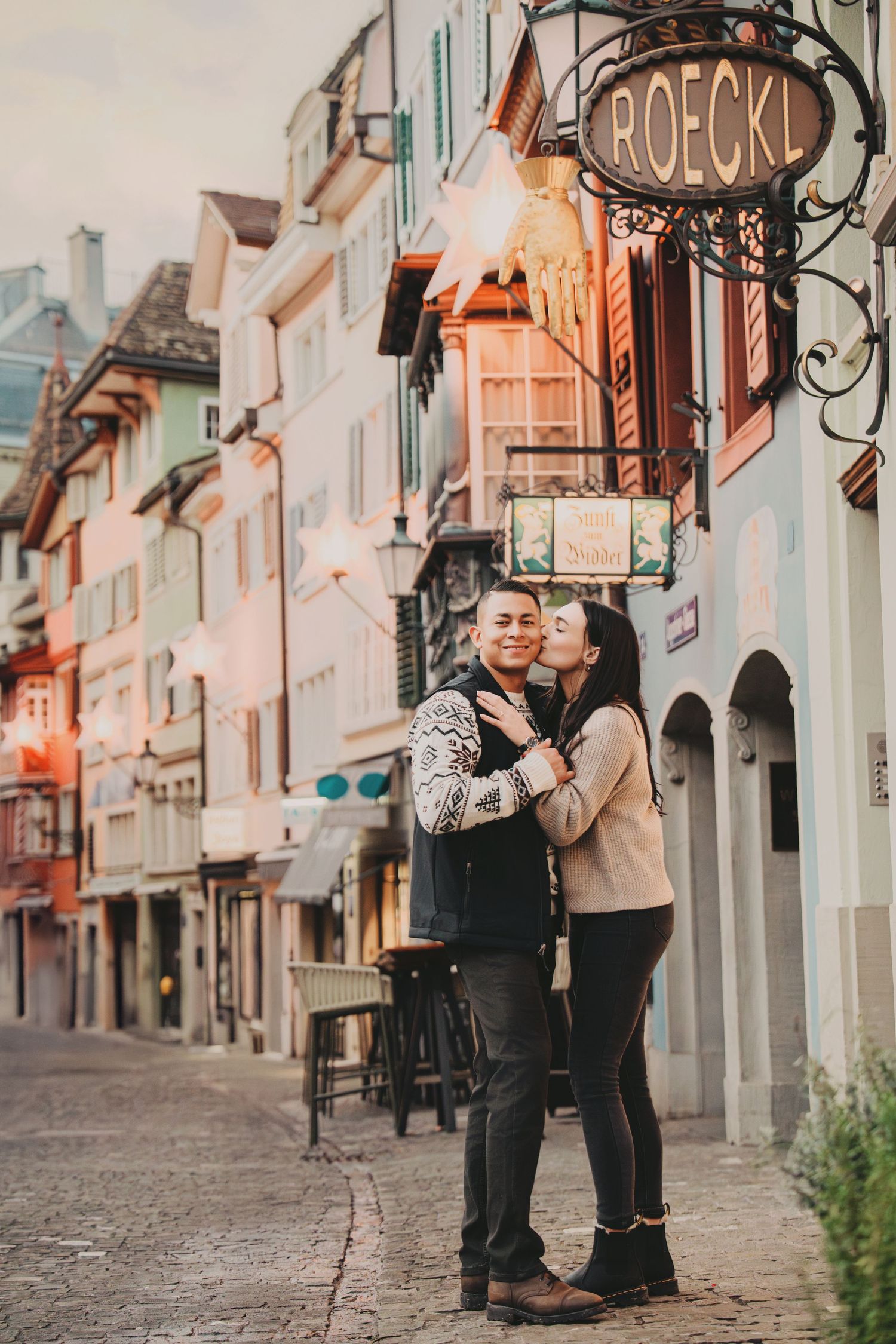 Woman kissing man on cheek while standing on a European cobblestone street at dusk.