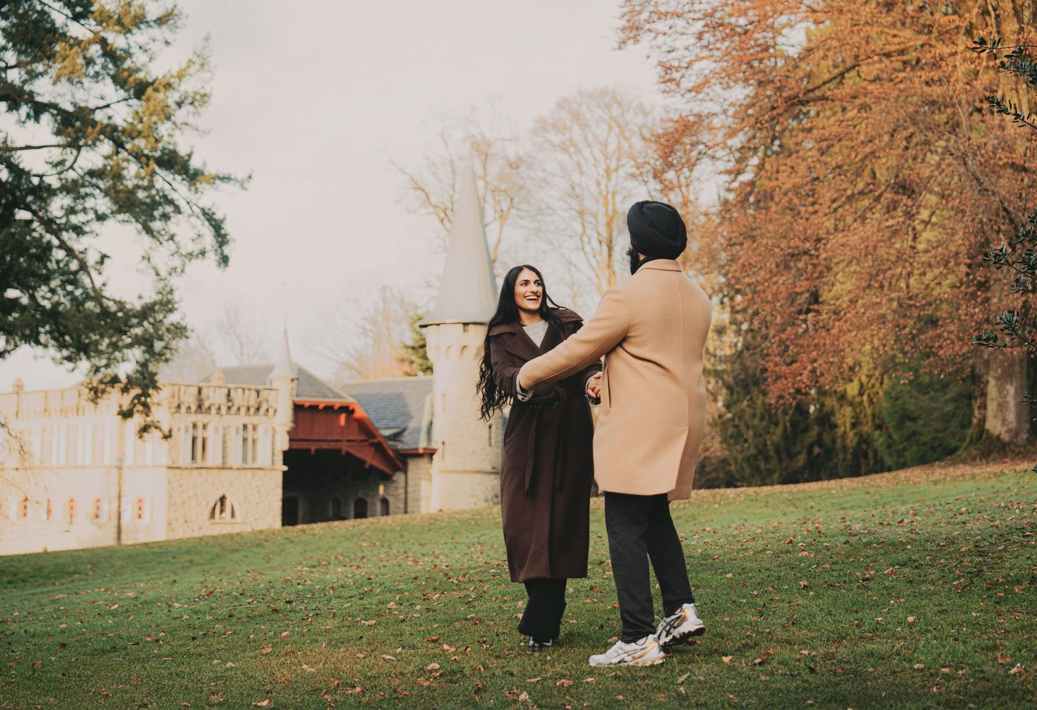 Couple holding hands and dancing playfully on a green lawn with autumn trees behind them.