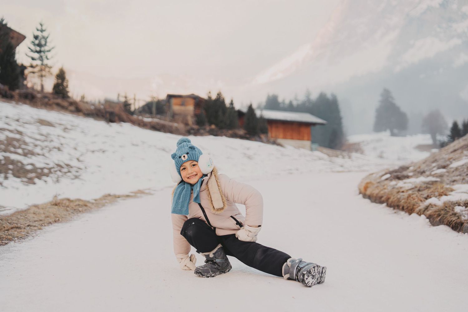 Young child in blue hat and scarf sitting playfully on snowy alpine road with mountains in background.