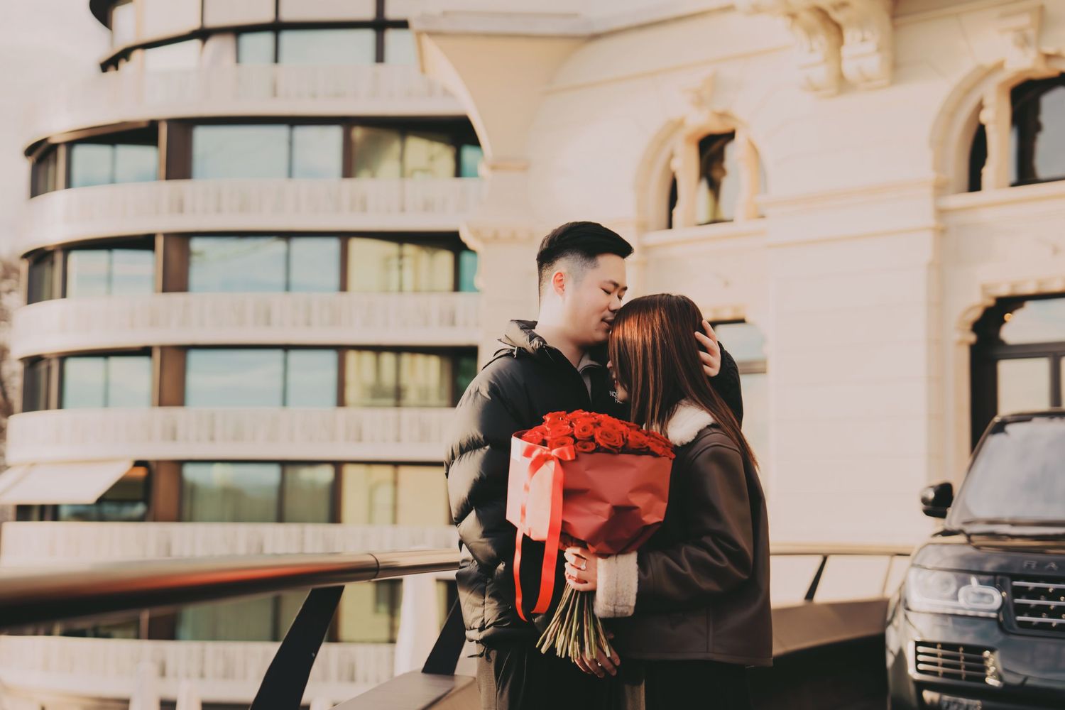Romantic couple sharing a kiss while holding a bouquet of red roses outside a luxury hotel.