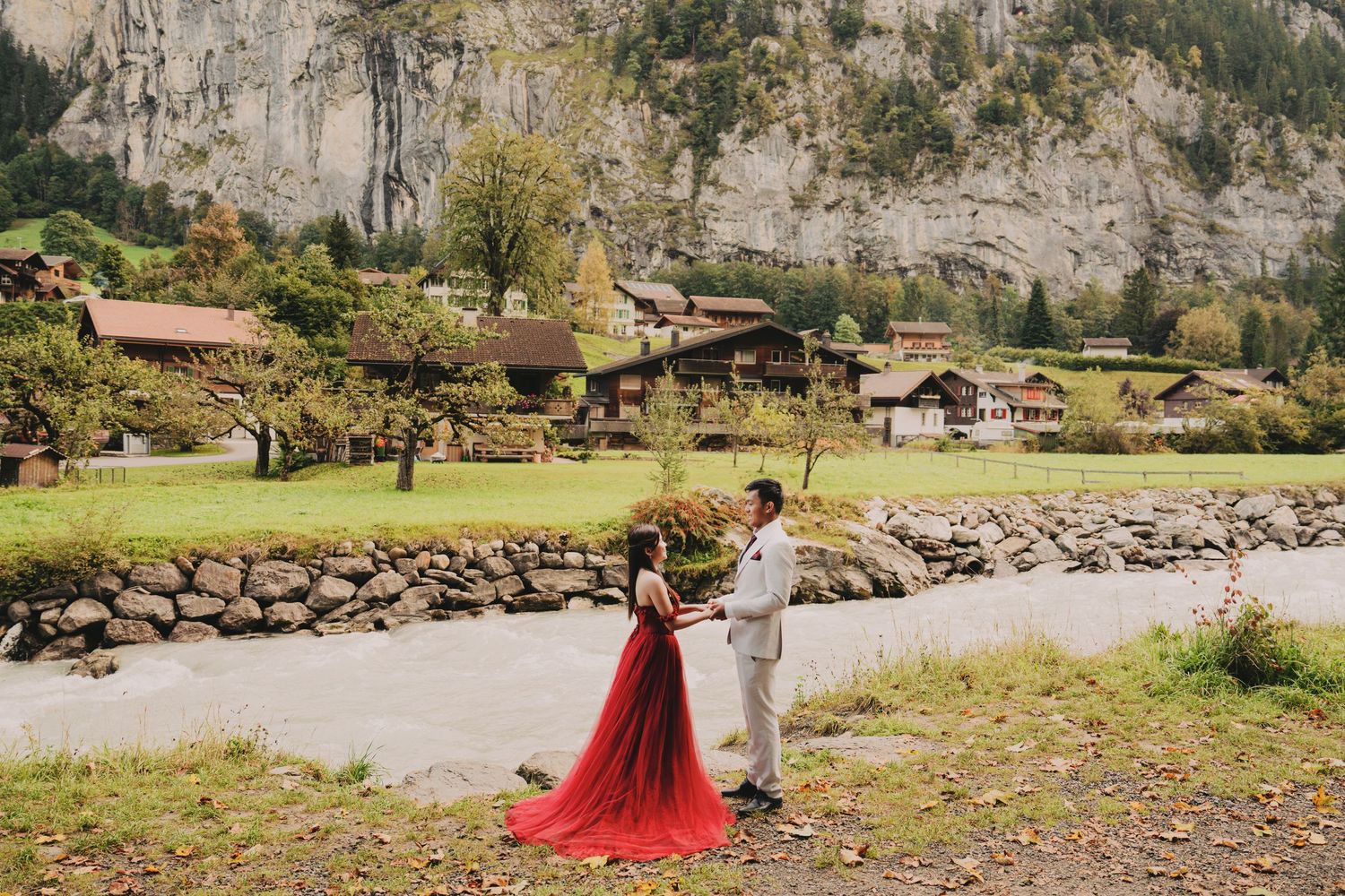 Couple dancing in red bridal gown beside river in charming Swiss mountain village setting.