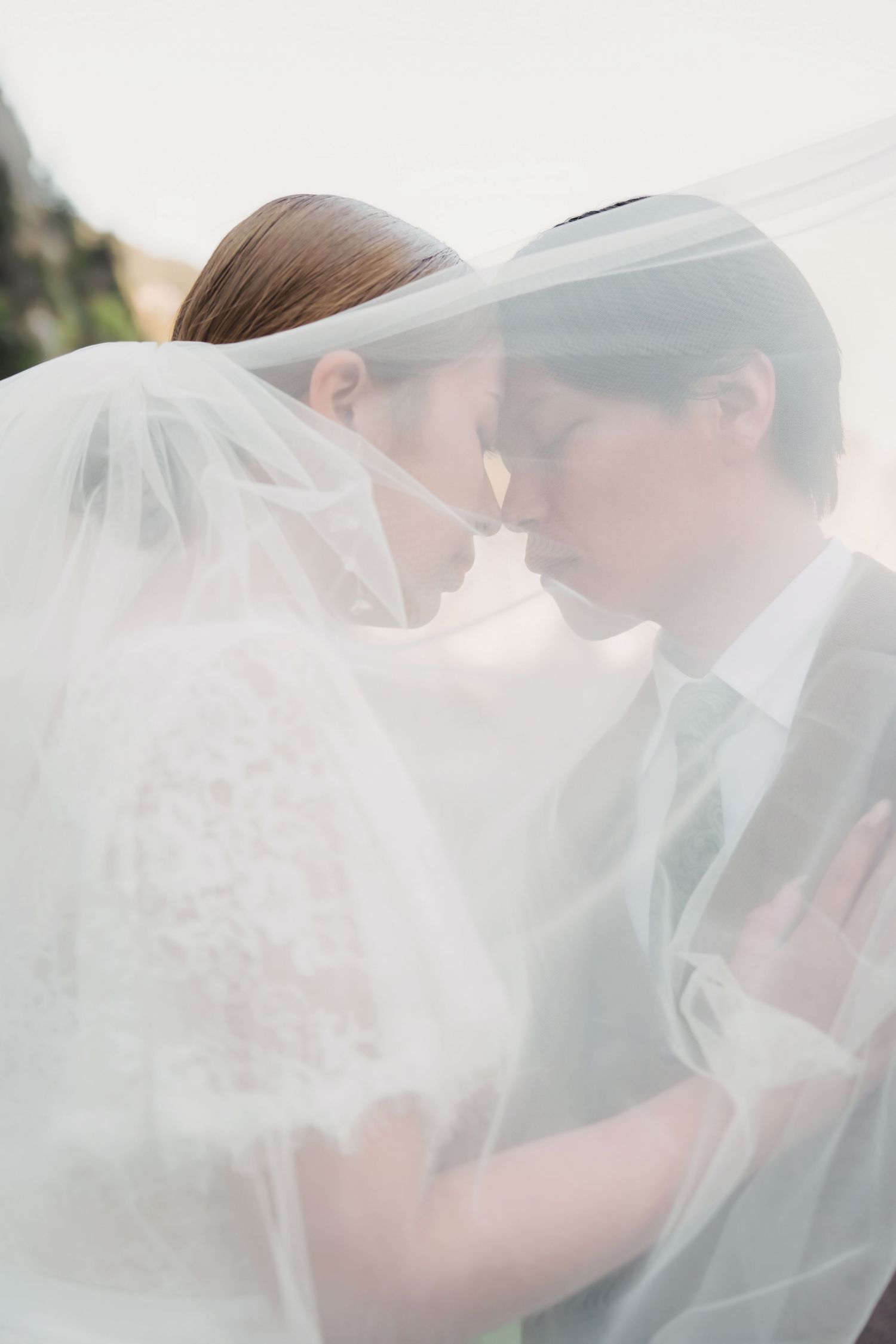 Wedding couple tenderly touching foreheads beneath a flowing white bridal veil in a soft romantic portrait.
