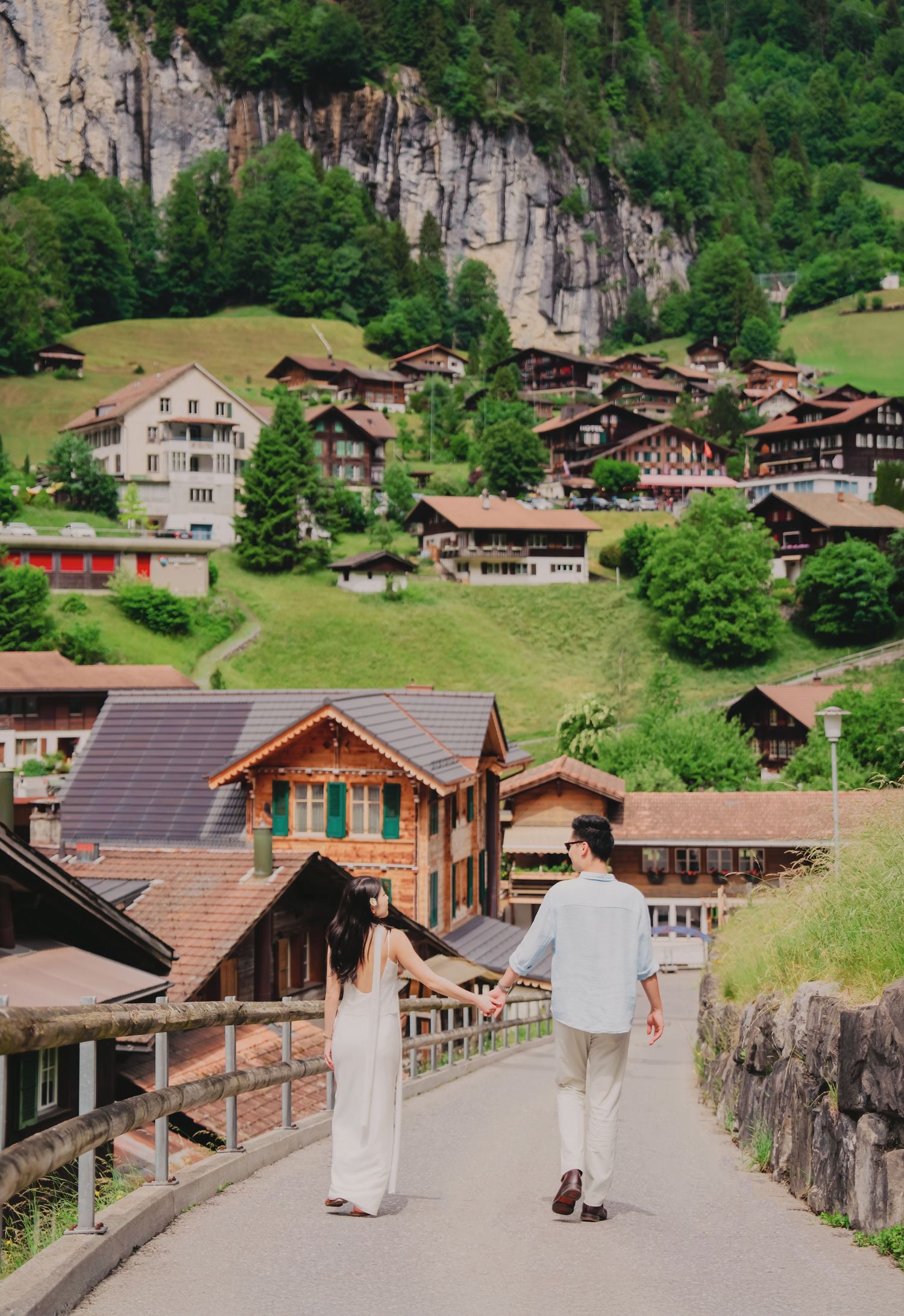 Couple holds hands walking toward charming Swiss alpine village with traditional wooden chalets on hillside.