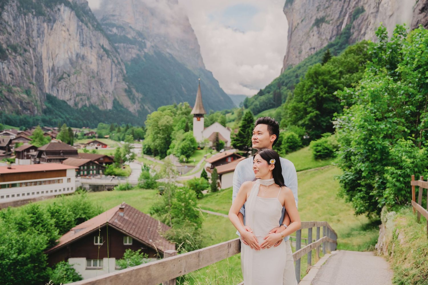 Couple embracing on a bridge overlooking a charming Swiss village with a church and mountain valley.