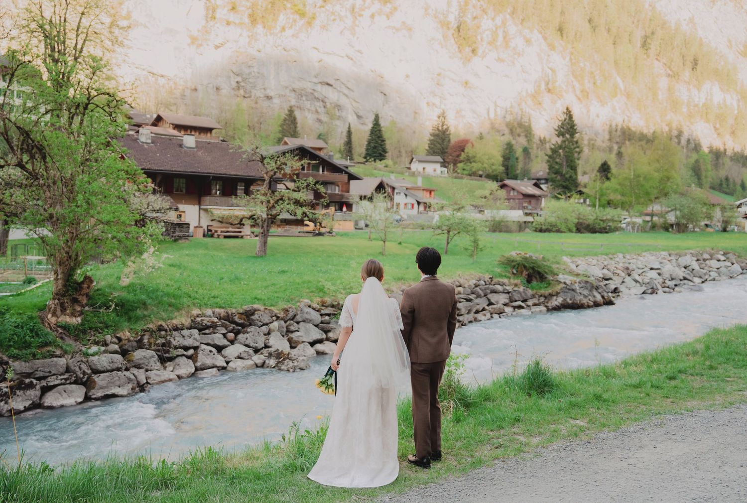 Wedding couple viewed from behind admiring a picturesque Swiss alpine village along a flowing river.