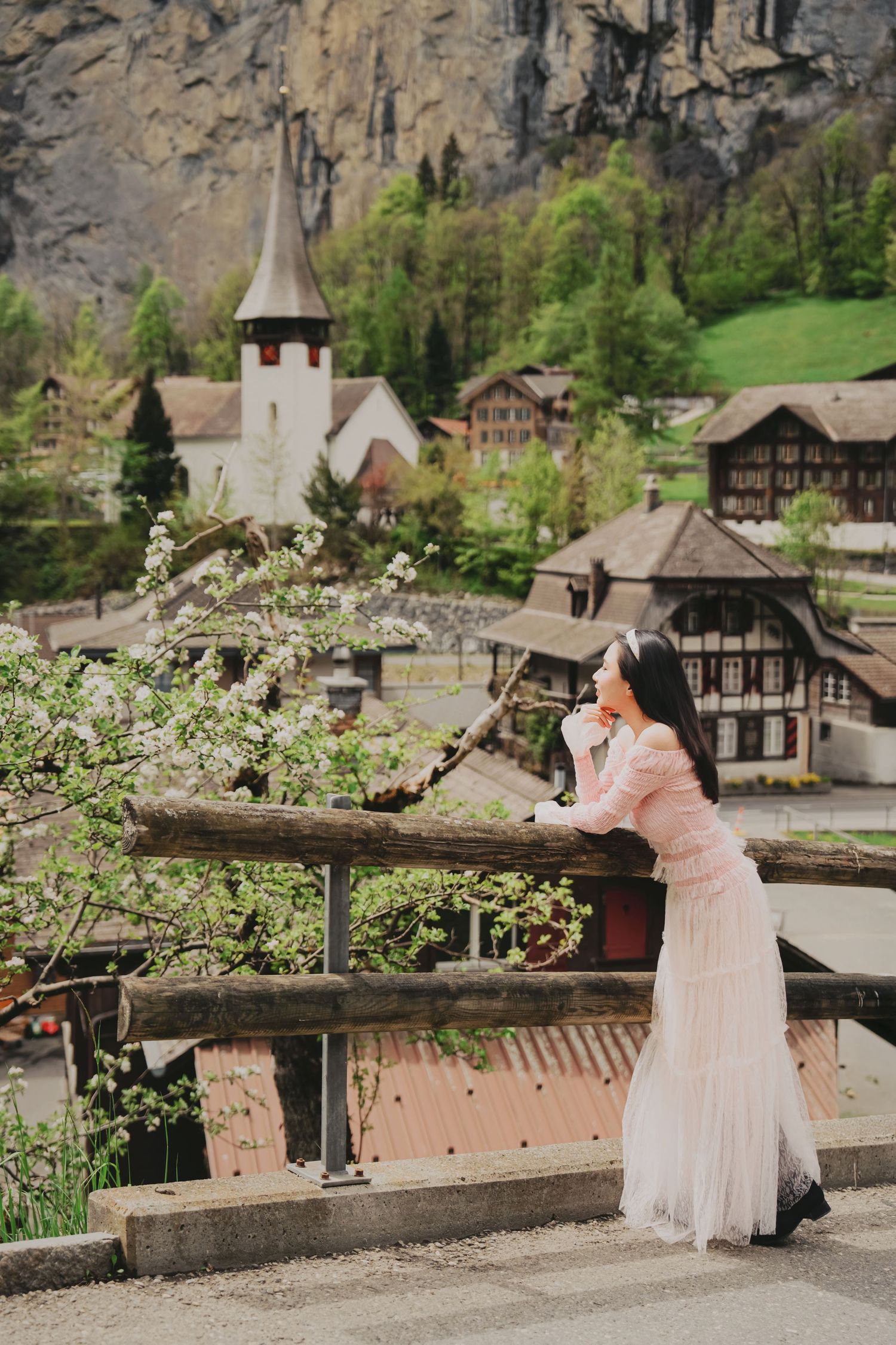 Woman in pink dress leaning on wooden railing overlooking Lauterbrunnen village church and alpine scenery.